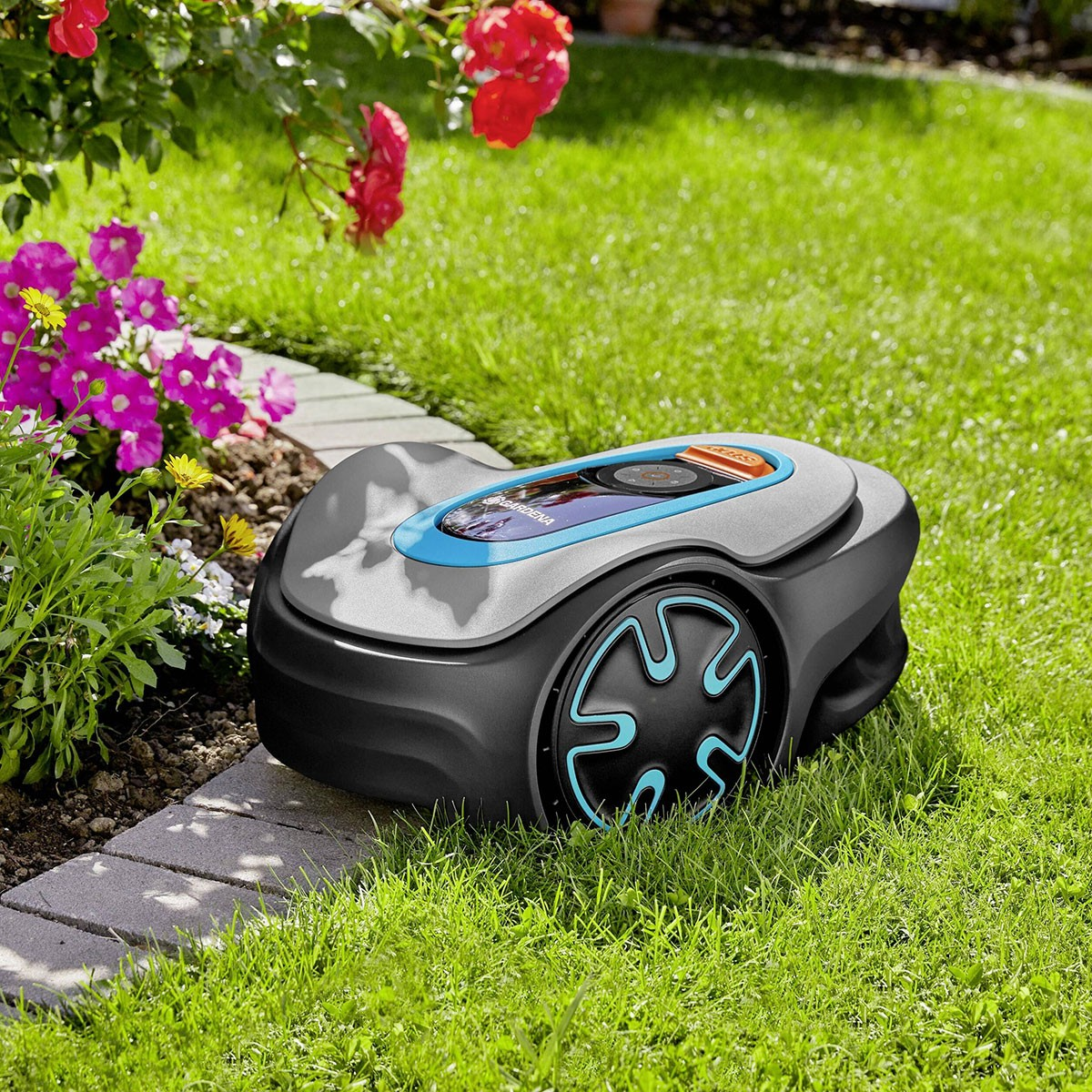 Mähroboter auf Gras, neben einem Blumenbeet mit rosa und roten Blüten.