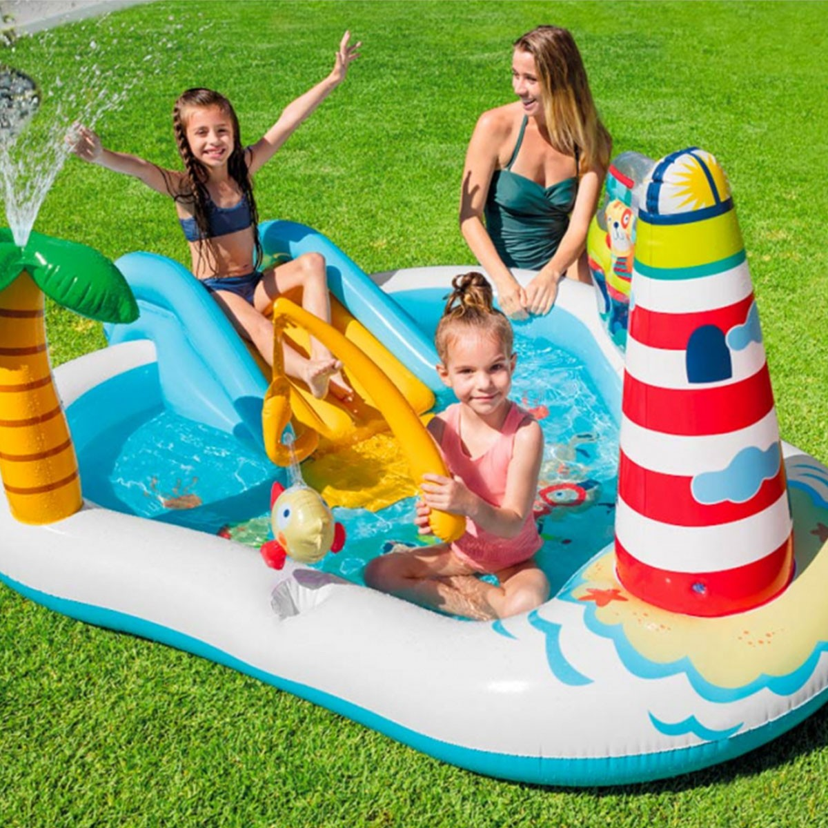 Bambini e una donna giocano in una piscina gonfiabile con scivolo, palma e faro.