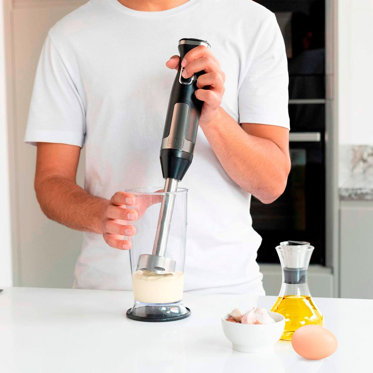 Osoba miksująca majonez w szklance blenderem ręcznym, składniki na stole.