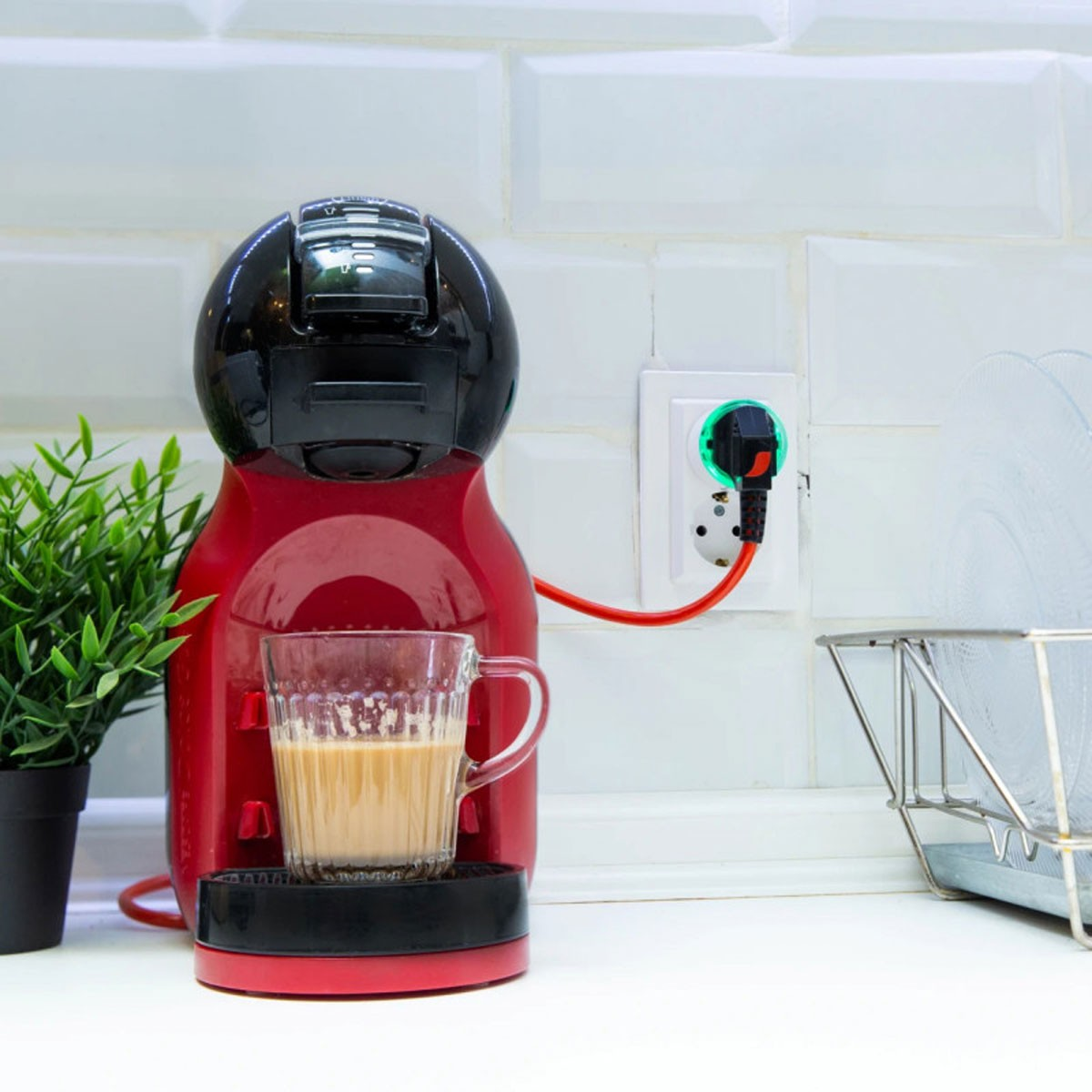Cafetera roja y negra con una taza. Un enchufe está en la pared. Fondo blanco.