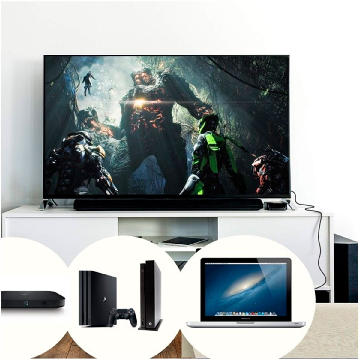 Un televisor muestra una escena de videojuego. Debajo consolas y un portátil. El fondo es una pared blanca.