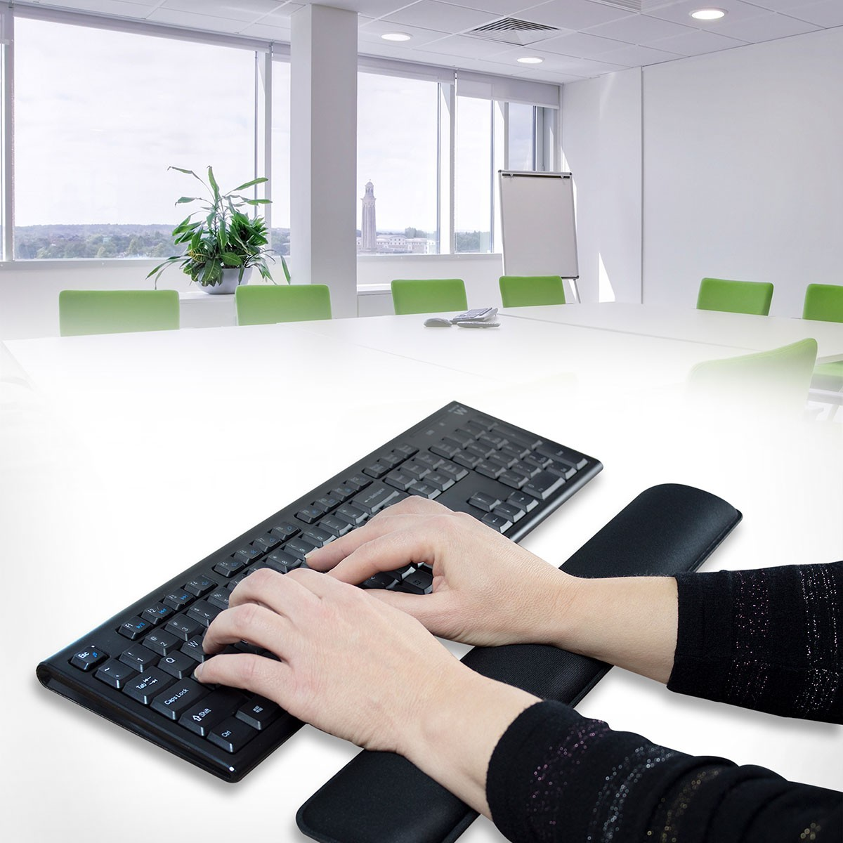 Manos escribiendo en un teclado negro con reposamuñecas en un entorno de oficina brillante.