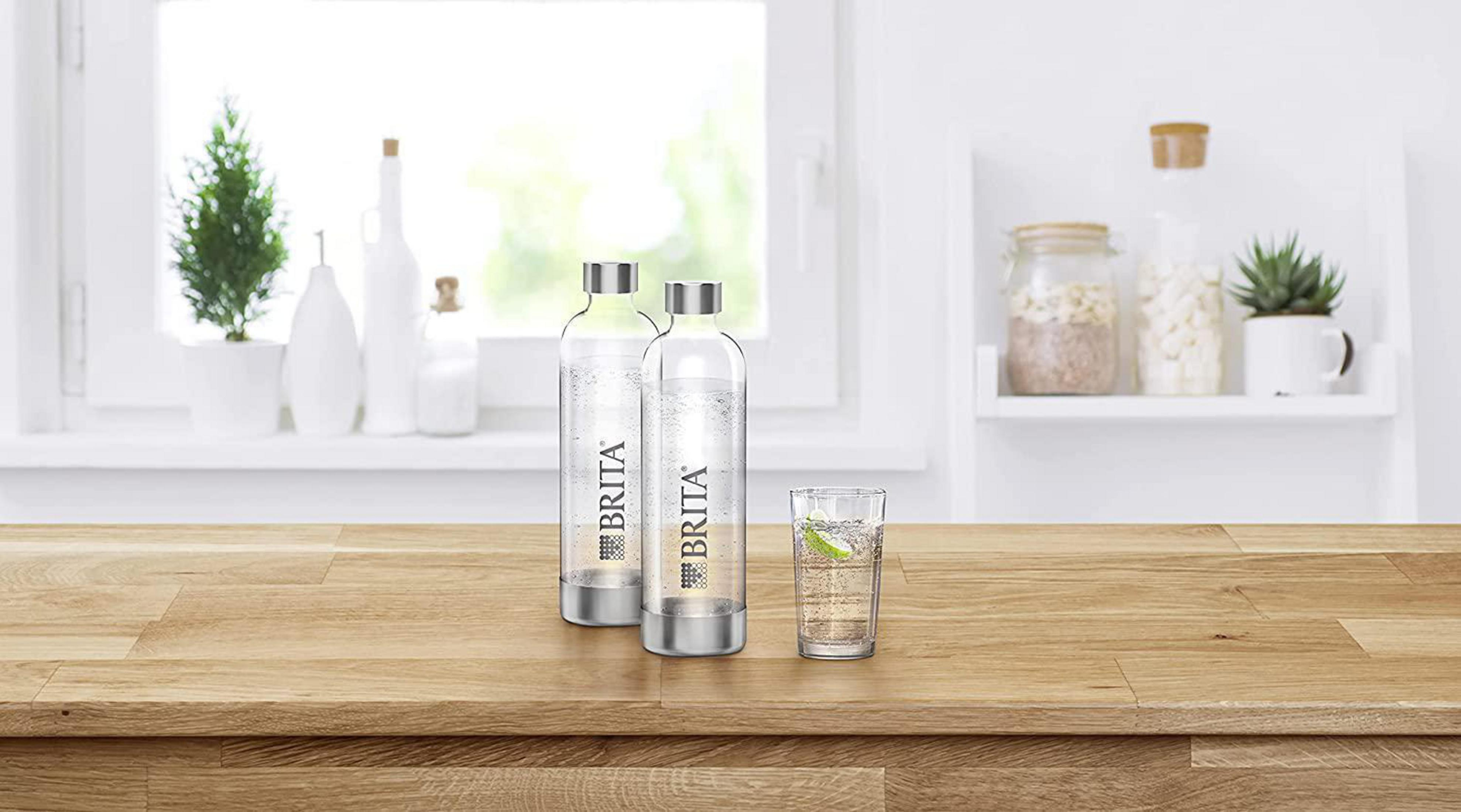 Zwei Glas-Brita-Wasserflaschen und ein Glas Wasser auf einem Holztisch. Regal und Fenster im Hintergrund.
