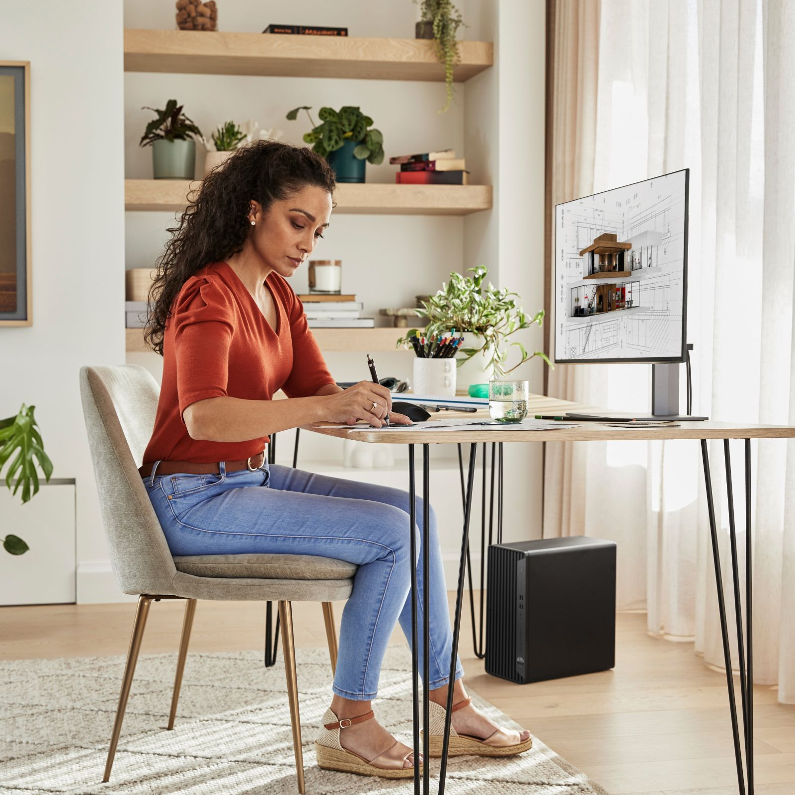 Frau schreibt an einem Schreibtisch mit Computer, der Hauspläne zeigt. Pflanzen im Hintergrund.