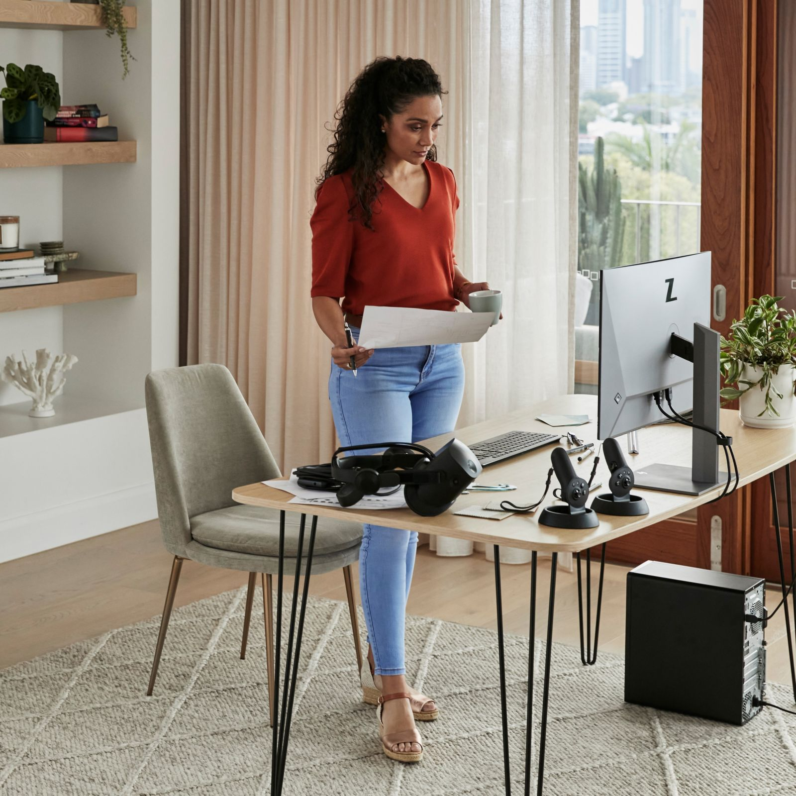 Frau steht an einem Schreibtisch, arbeitet mit Computer, VR-Headset und Papieren.