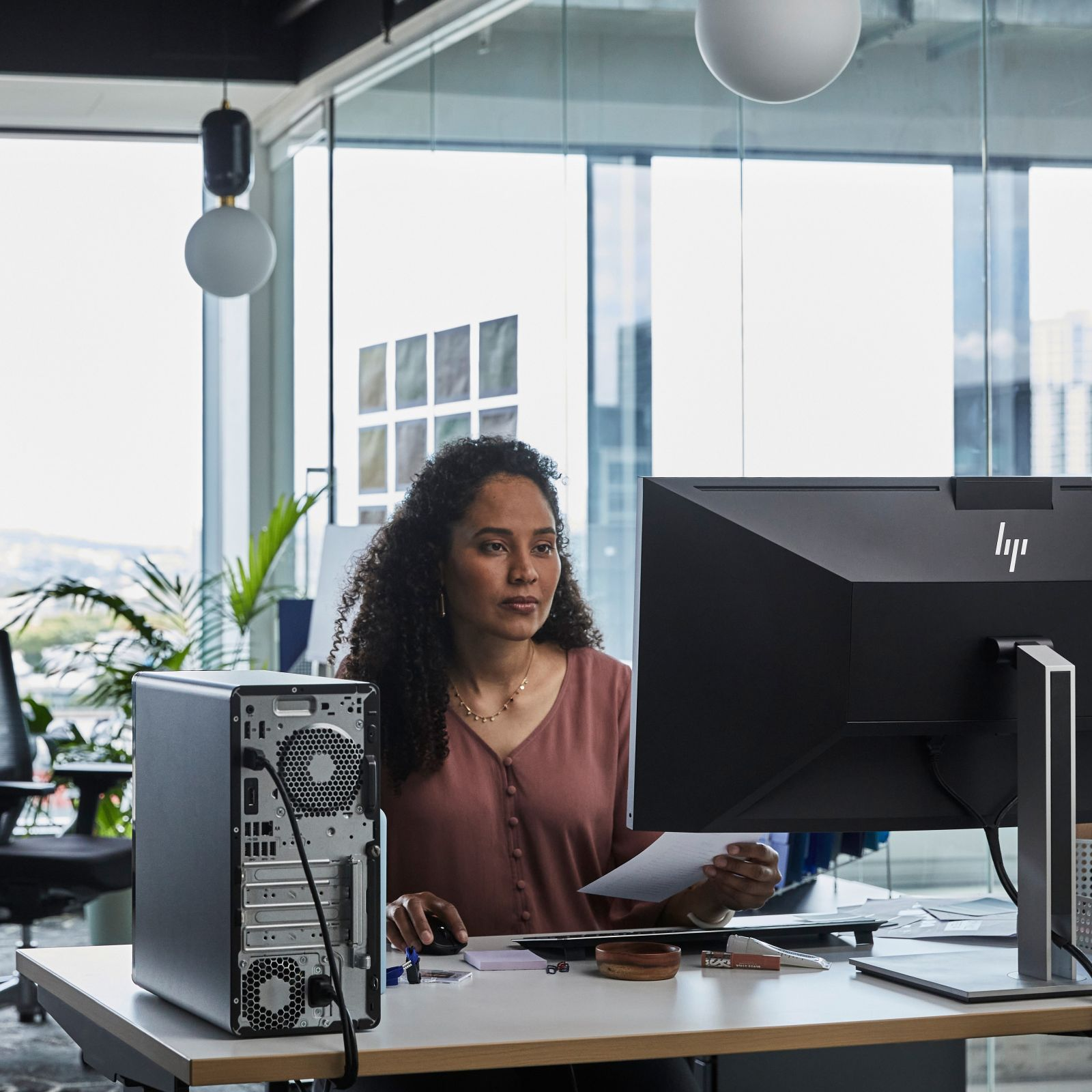 Frau am Schreibtisch, die ein Papier betrachtet, mit Monitor und Computergehäuse links.