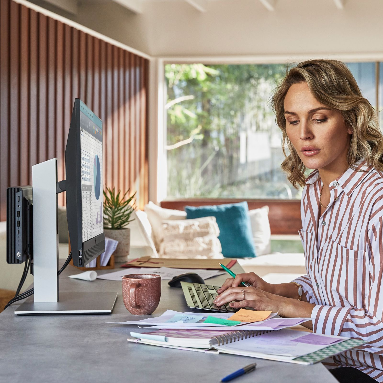 Kobieta przy biurku pracuje na komputerze i monitorze. Obecny jest kubek i roślina.