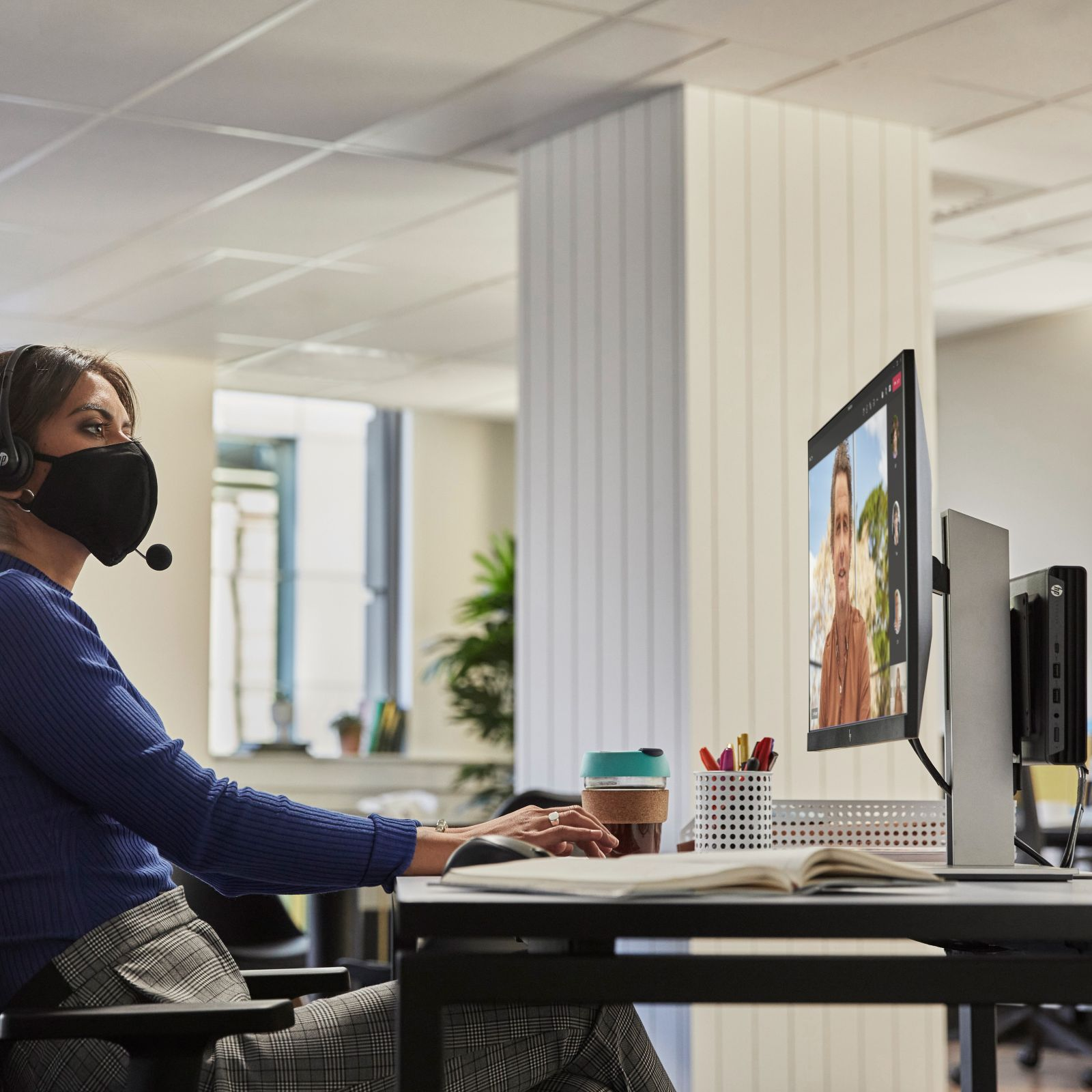 Kobieta w masce podczas rozmowy wideo, z monitorem i biurkiem z przedmiotami.
