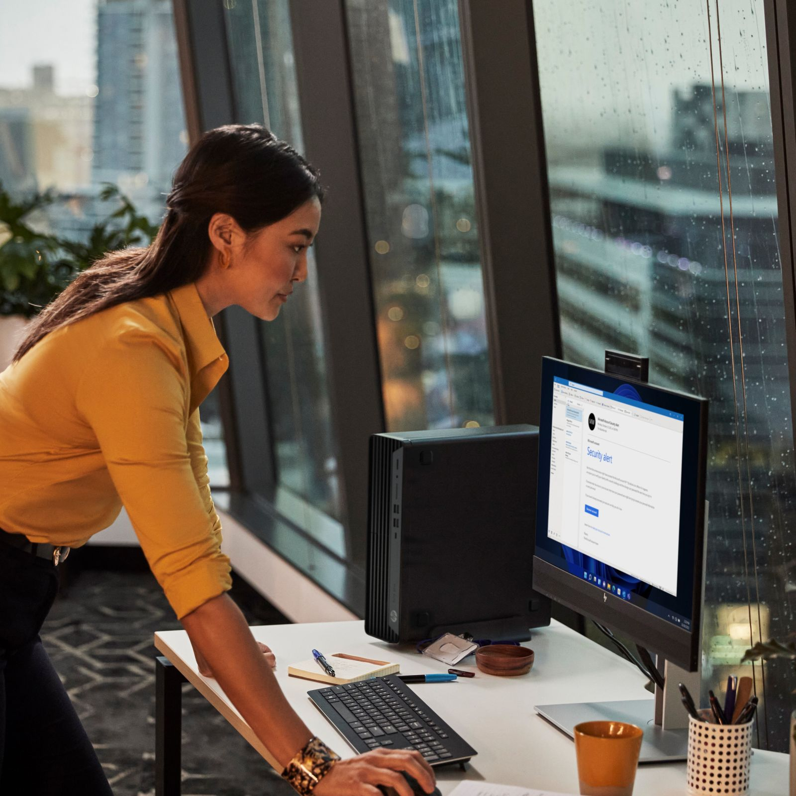 Eine Frau in gelbem Hemd arbeitet an einem Schreibtisch mit Computer und Monitor.