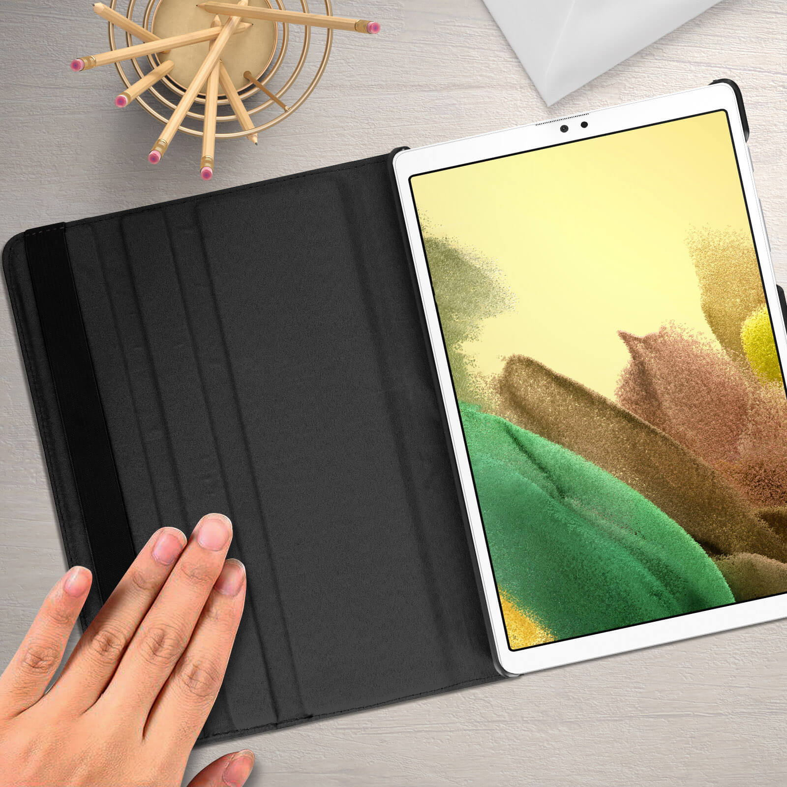 Tablet su una custodia nera, con una mano vicino. Lo sfondo include matite in un supporto.