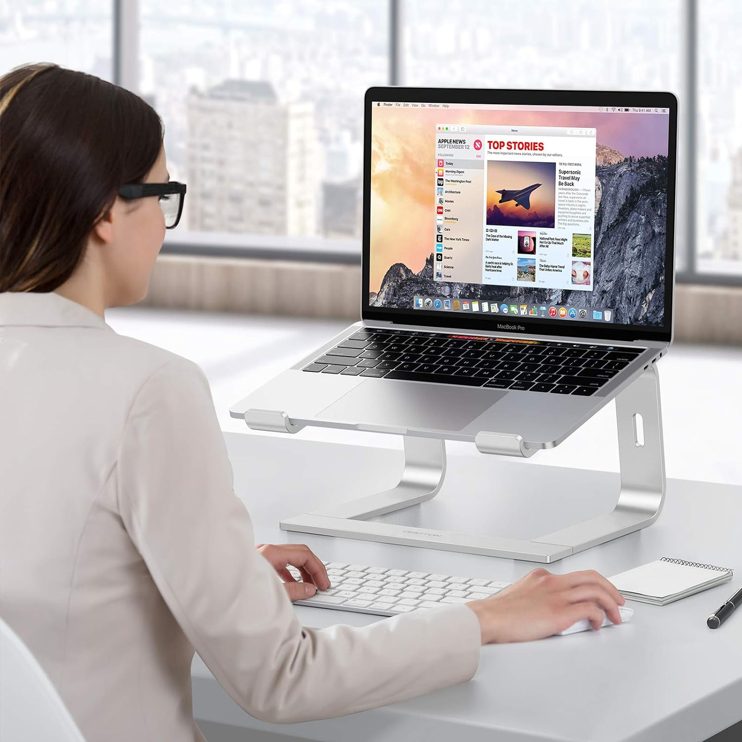 Frau benutzt Laptop auf Ständer, Tastatur und Maus sichtbar. Der Hintergrund ist ein Schreibtisch.