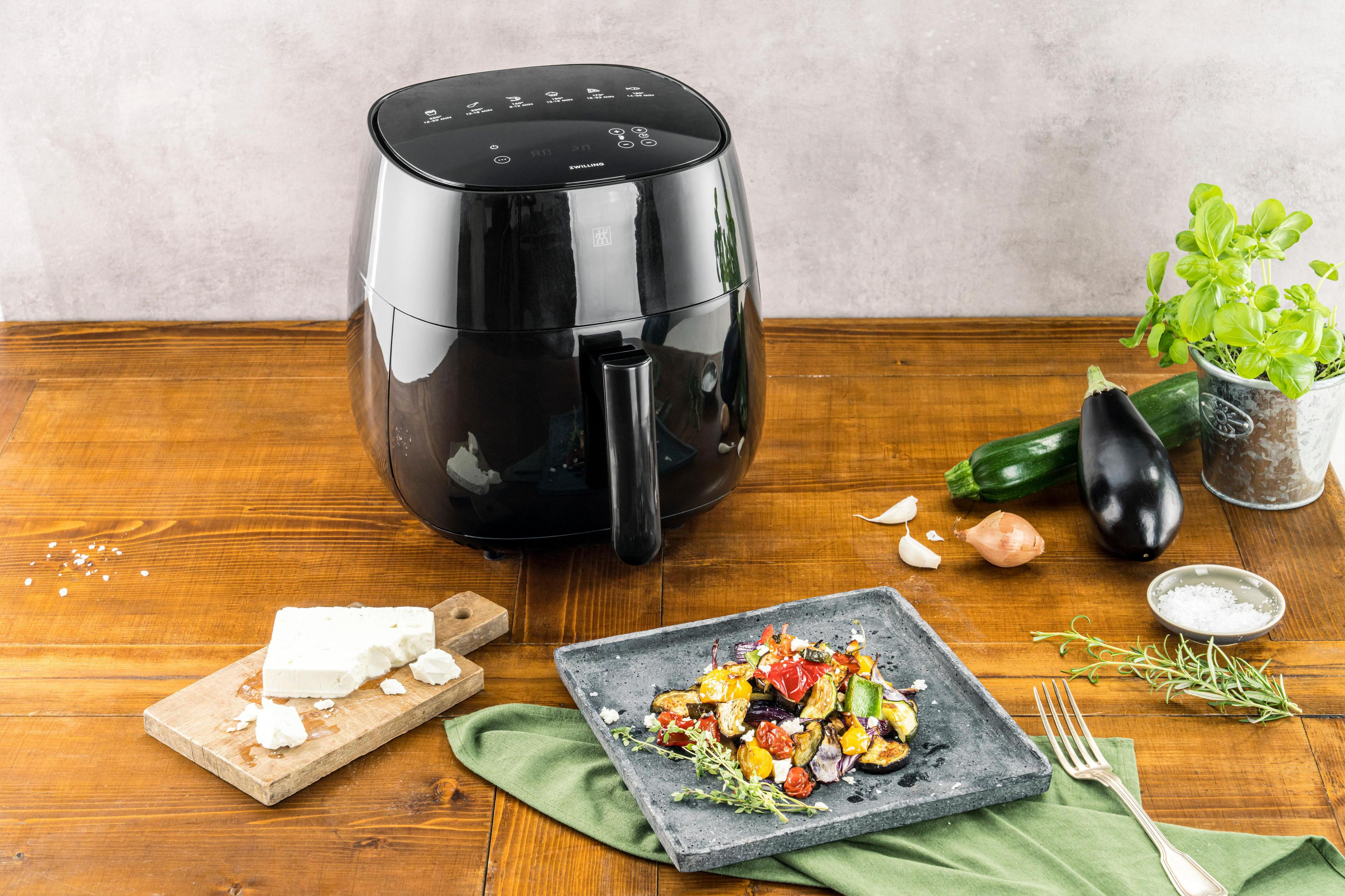 Czarna frytownica, gotowane jedzenie, warzywa i składniki na drewnianym stole. Tło to ściana.