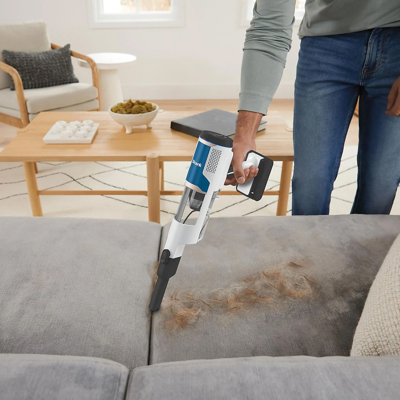 Person saugt ein graues Sofa mit einem weiß-blauen Staubsauger, der Tierhaare entfernt.