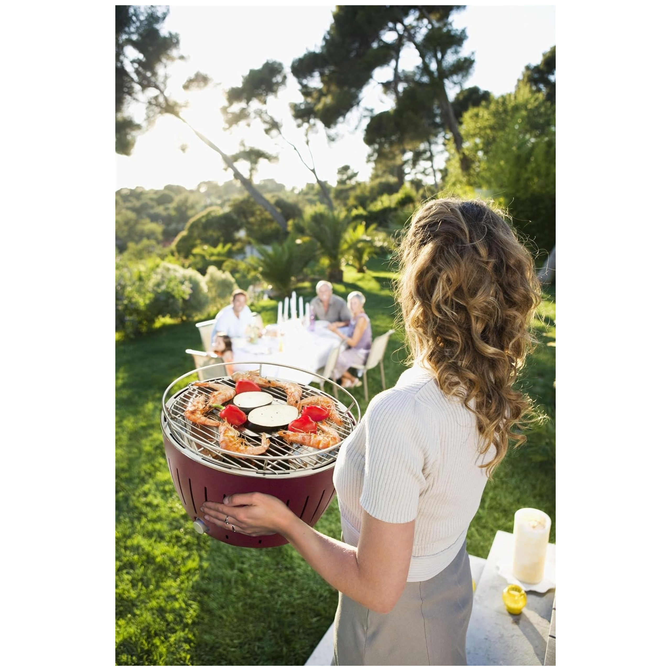 Kobieta trzymająca grilla z jedzeniem, w tle ludzie jedzący obiad, słonecznie na zewnątrz.