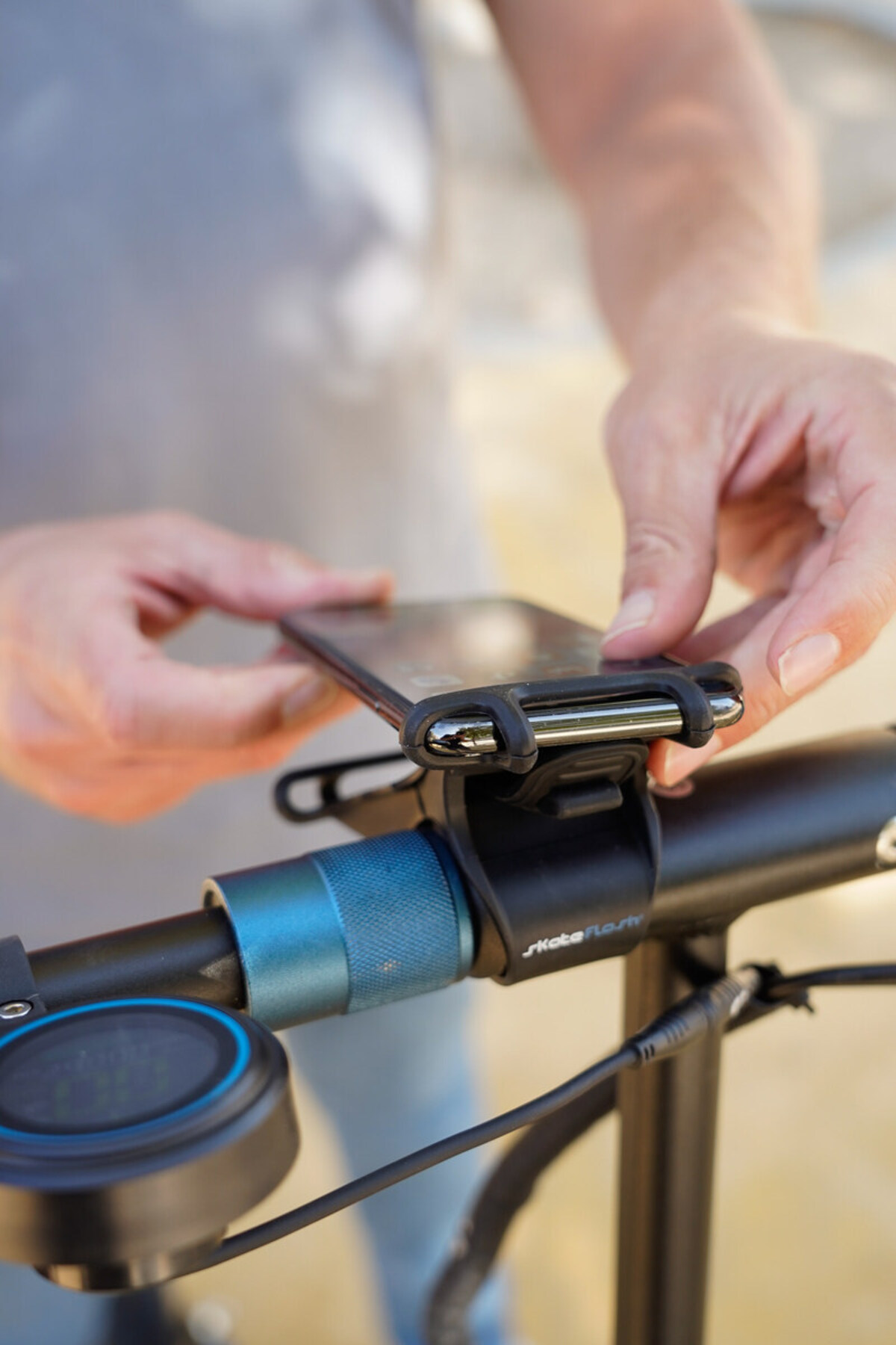 Las manos de una persona están uniendo un teléfono al manillar de un scooter. El scooter tiene un anillo azul y un mango negro.