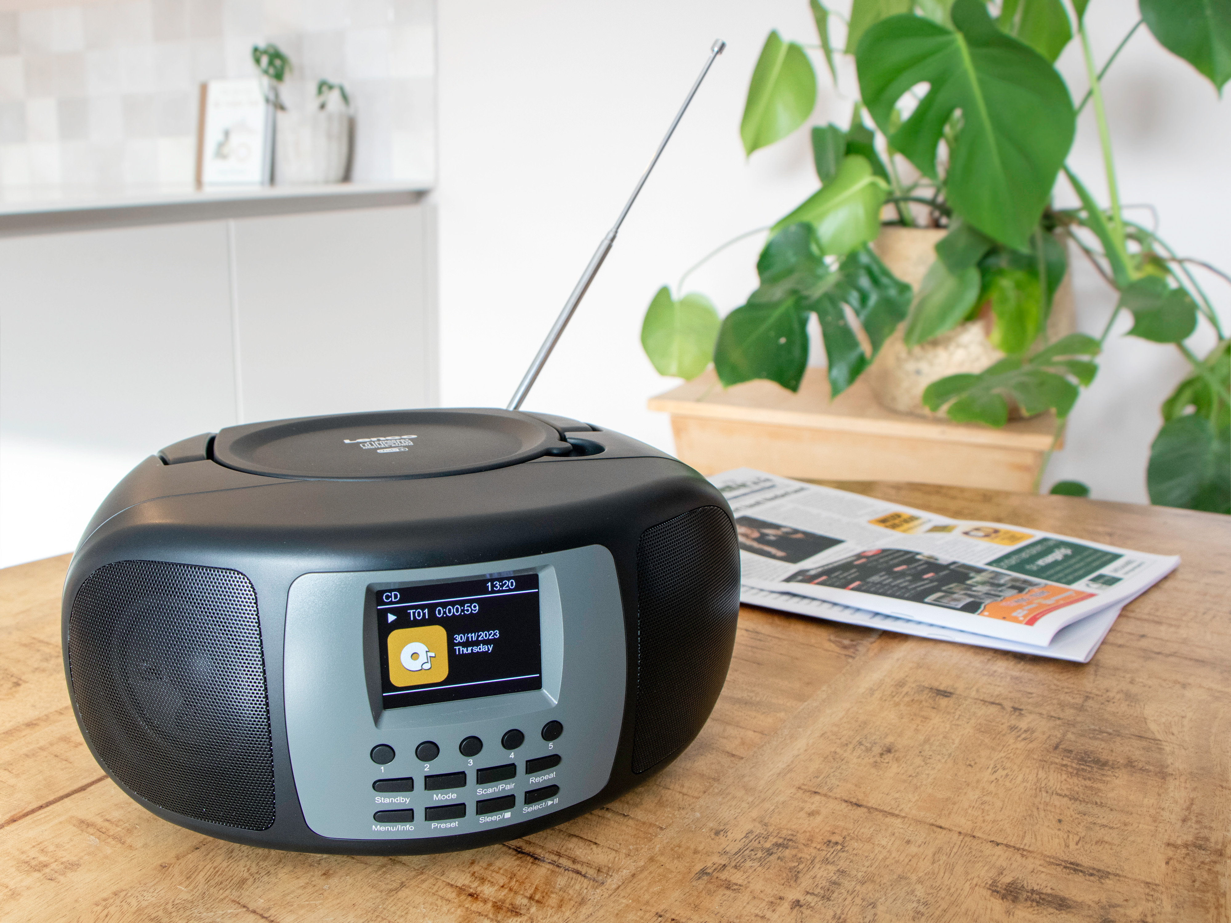 A black radio with a CD player on a wooden table, with a newspaper and a plant.