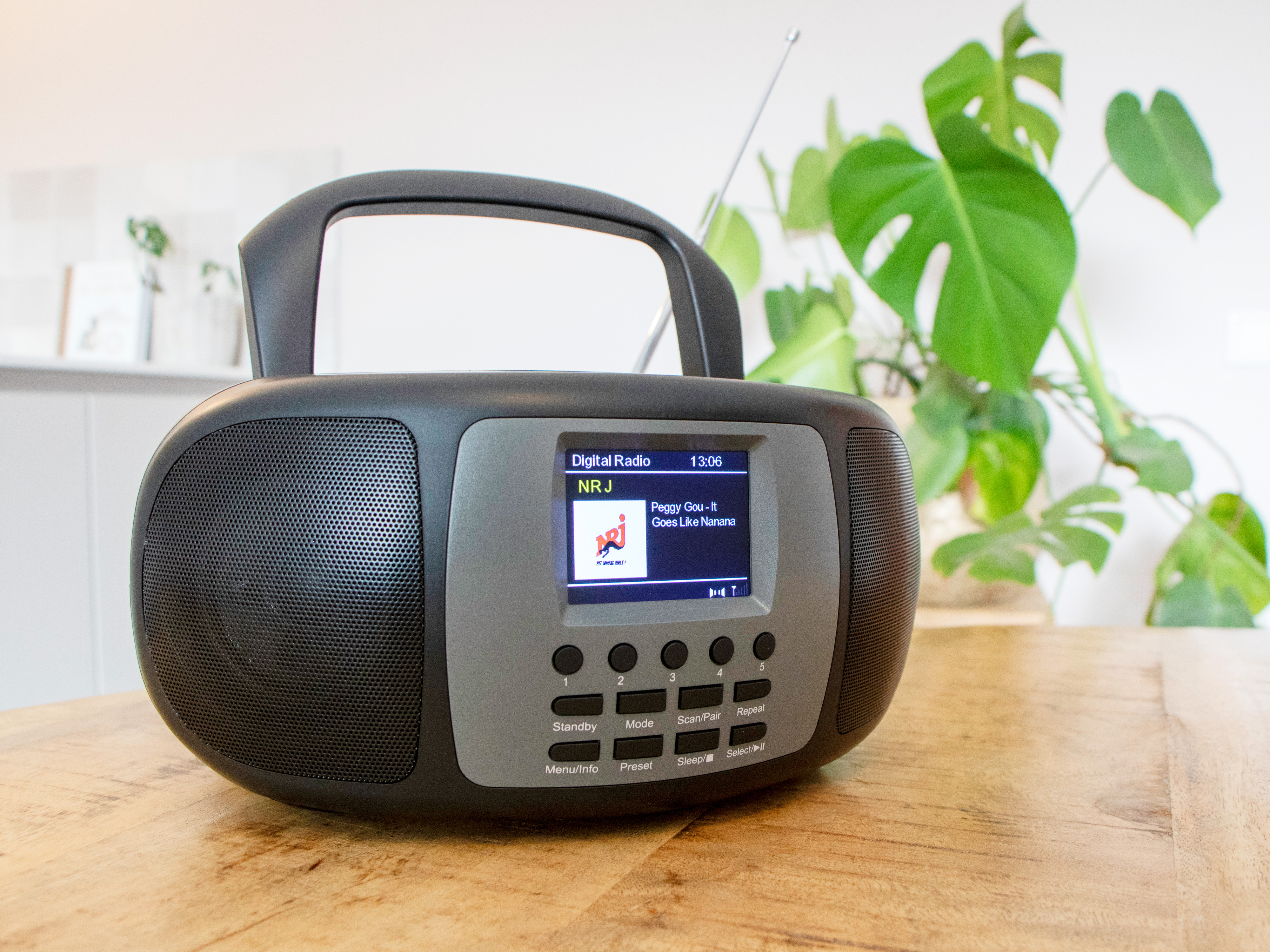 A black and gray radio with a digital display, showing a song title and time, on wood table.
