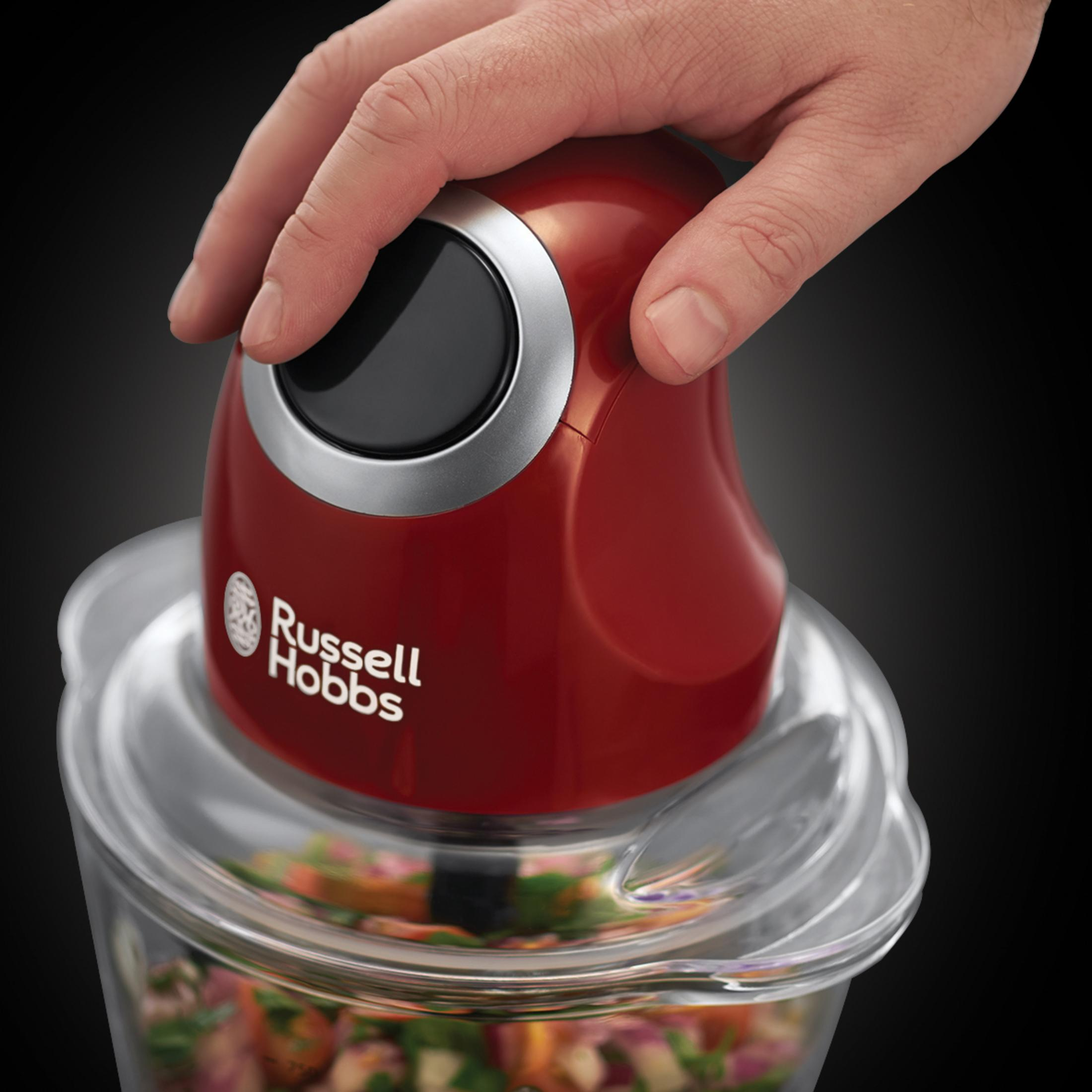 Eine Hand befindet sich über einem roten Zerkleinerer. Der Zerkleinerer steht auf einer weißen Oberfläche, mit Essen darin.