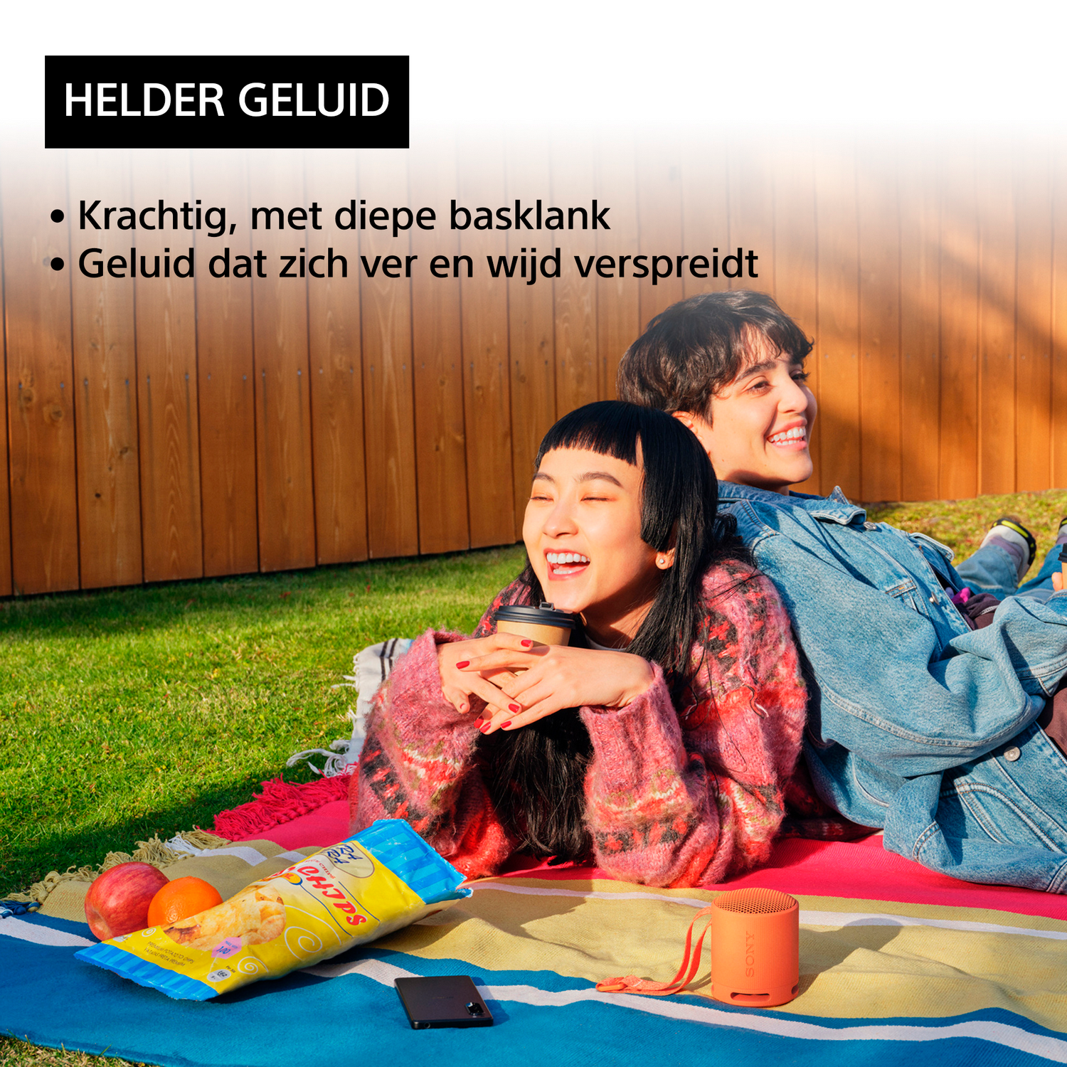 Dwie osoby uśmiechają się i śmieją na kocu z przekąską i głośnikiem w pobliżu, na słońcu.