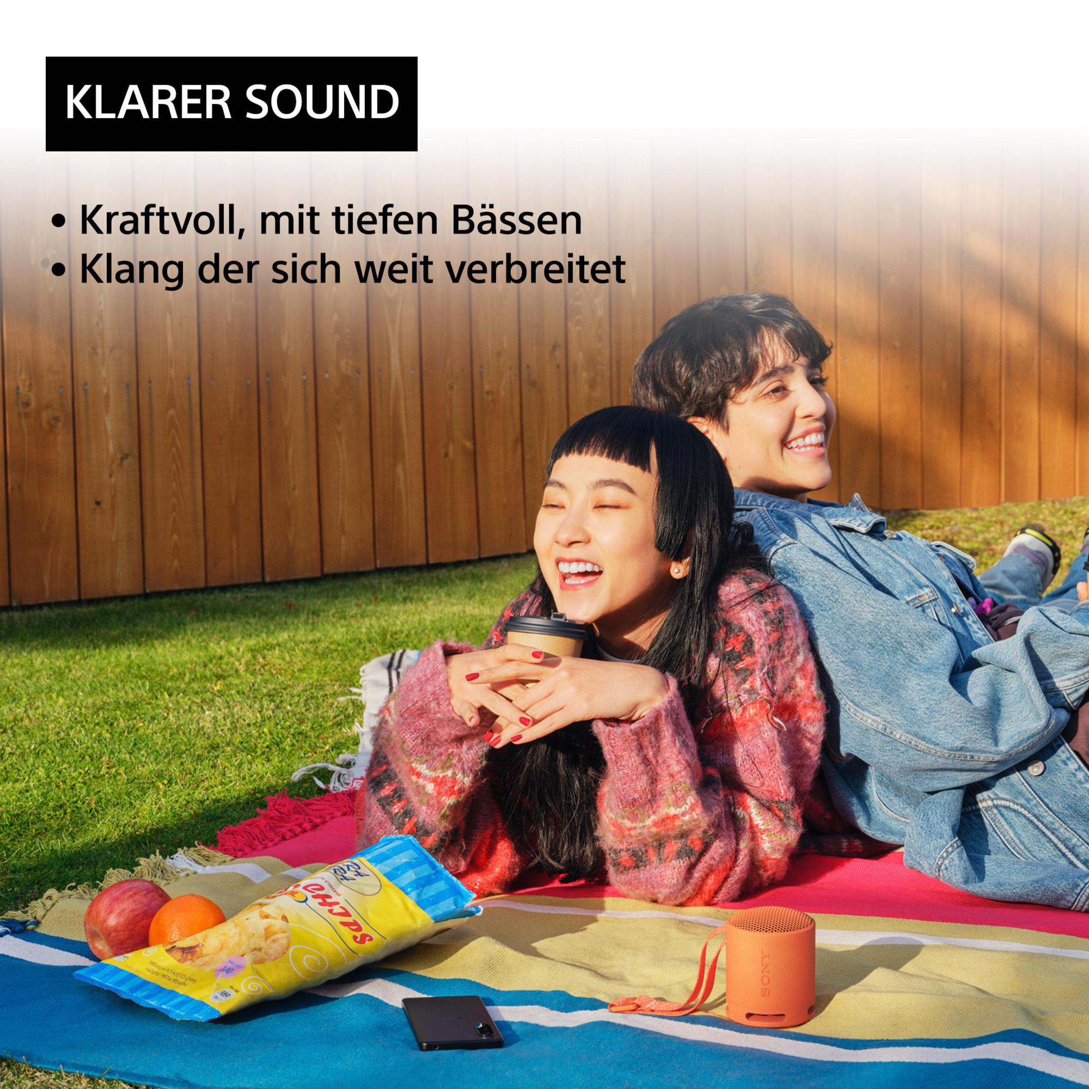 Dwie osoby uśmiechają się, siedząc na kocu z przekąskami, głośnikiem i czarnym telefonem.