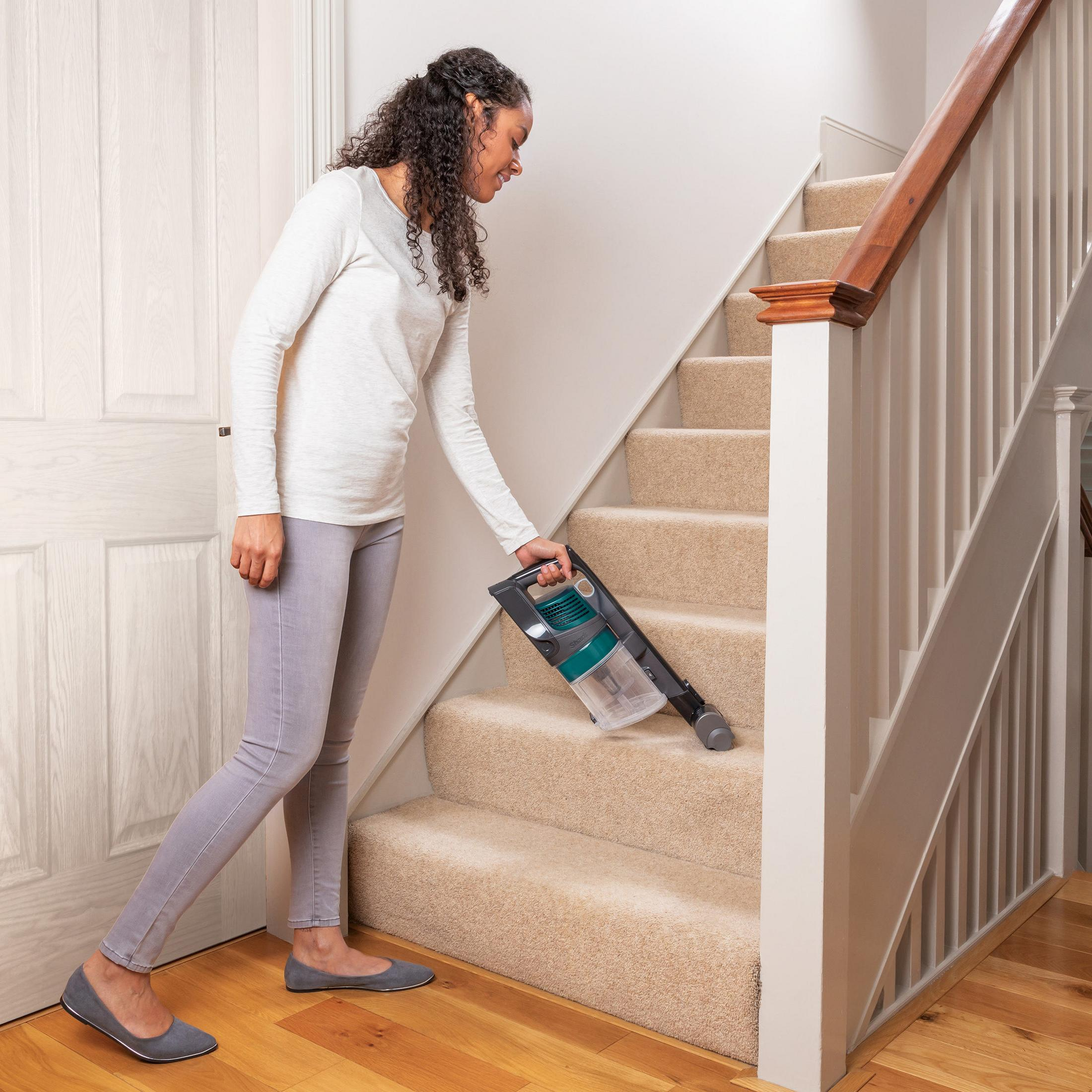 Mujer aspira escaleras con aspiradora de mano. Escaleras tienen alfombra beige y barandilla blanca.