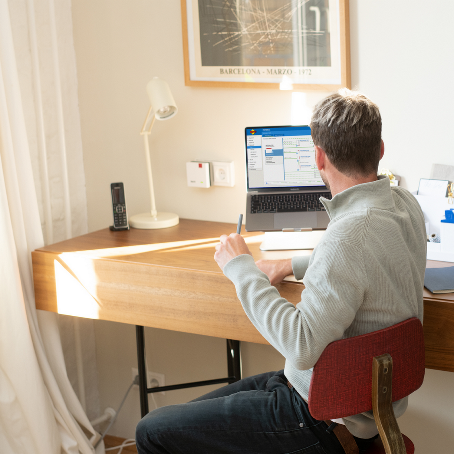 Mężczyzna siedzi przy drewnianym biurku, używając laptopa. W scenie widoczna jest lampa, telefon i plakat.