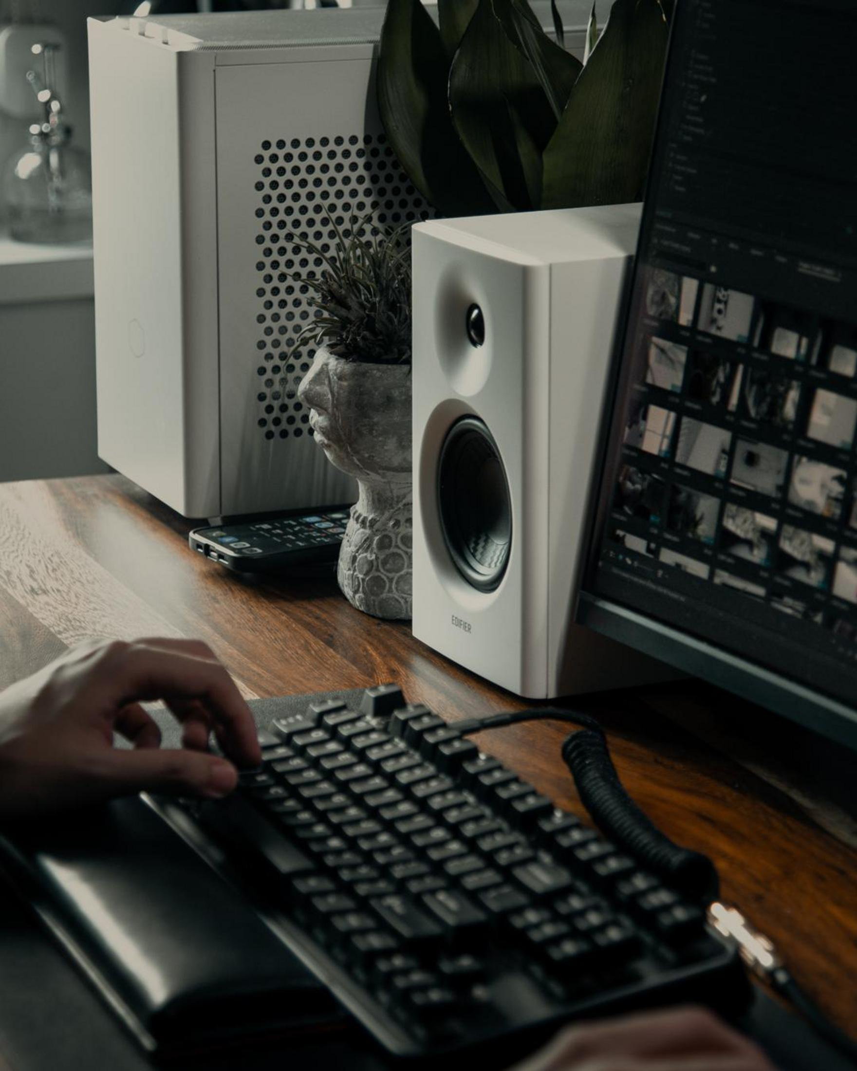 Biurko z komputerem, głośnikami, klawiaturą i rośliną doniczkową. Scena jest w pomieszczeniu. Oświetlenie jest słabe.