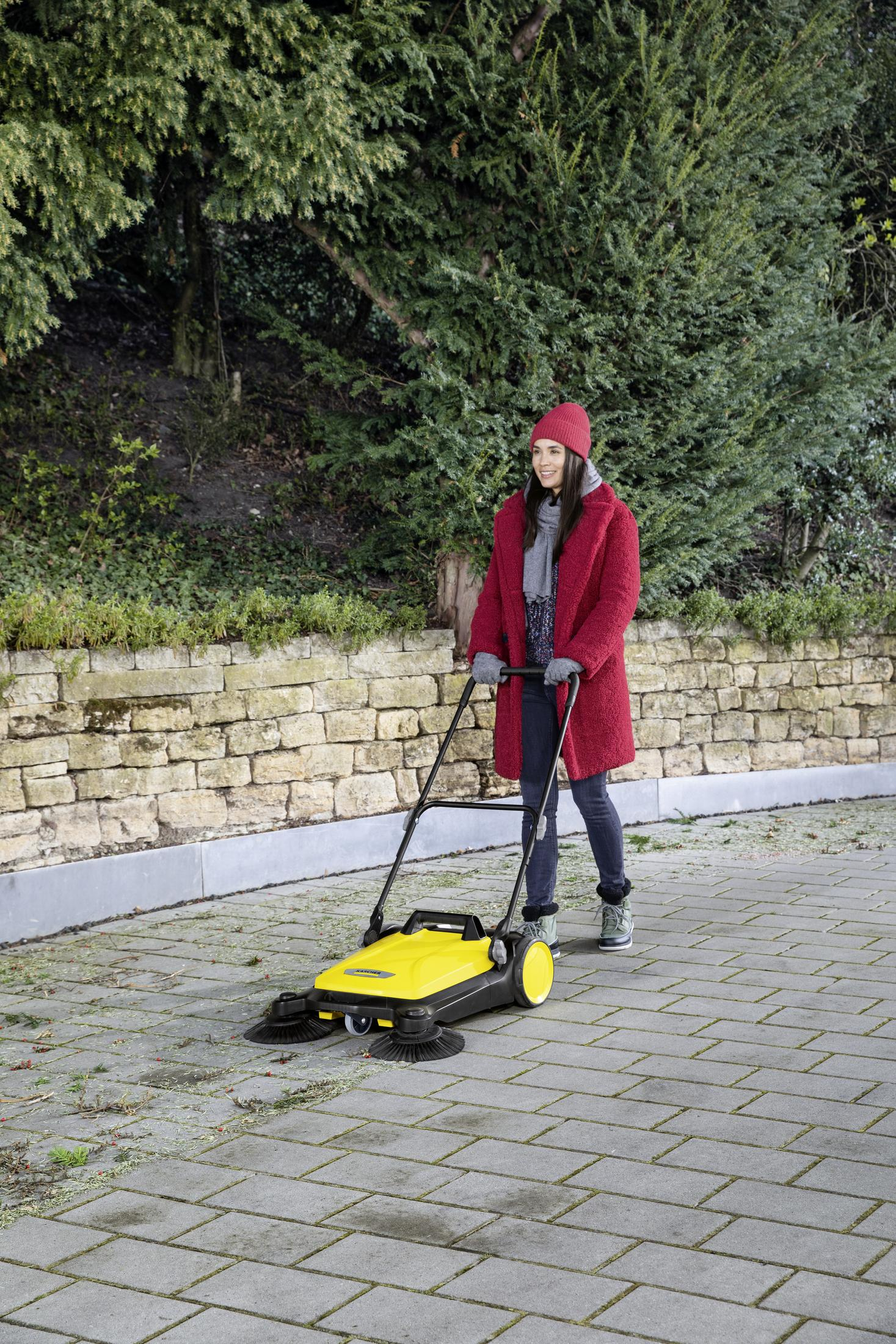 Kobieta w czerwonym płaszczu zamiata kamienną ścieżkę żółtą zamiatarką, za nią drzewa i mur.