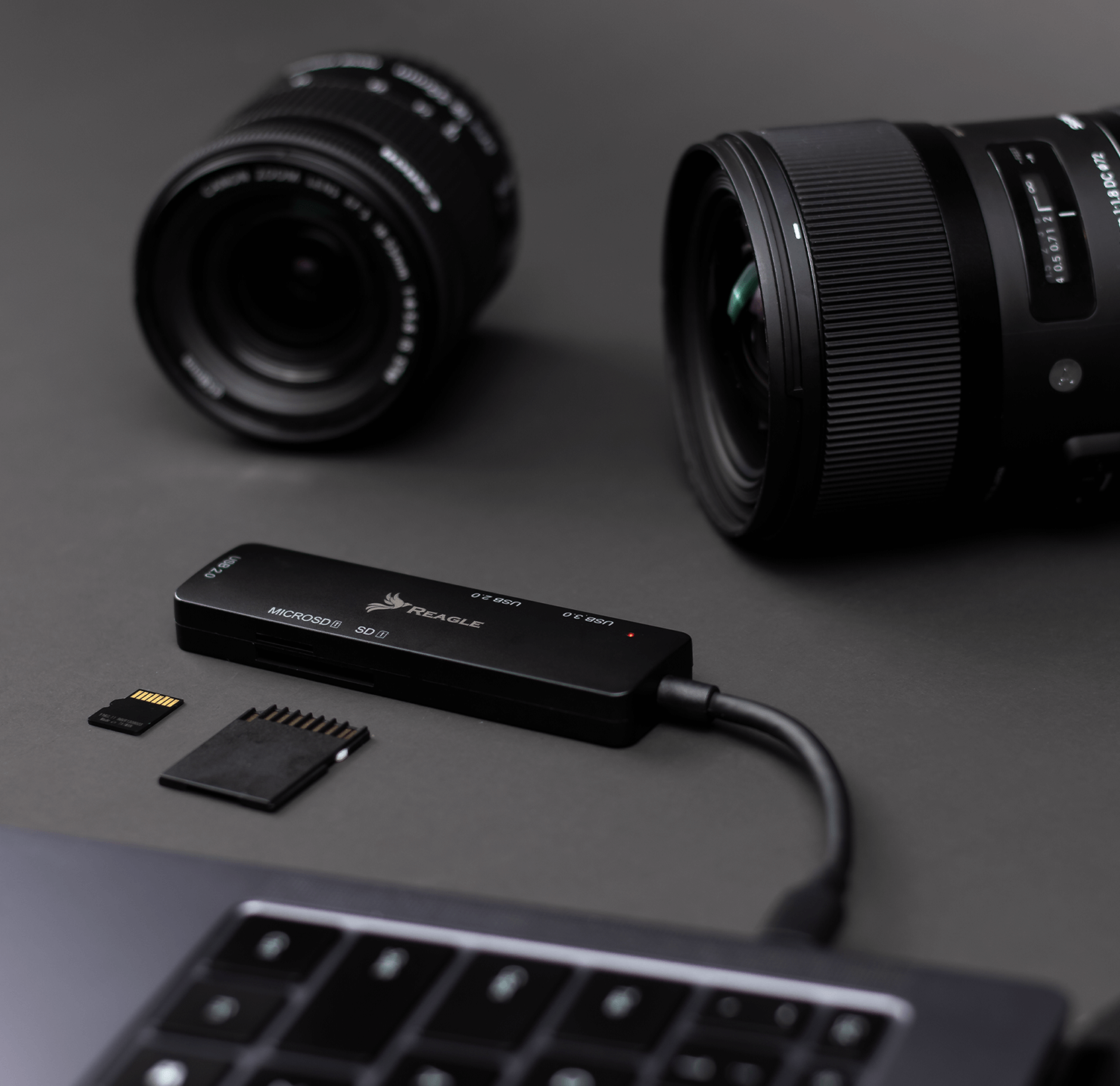 Schwarzes Kartenlesegerät mit SD- und MicroSD-Karten neben Canon- und Sigma-Objektiven auf dunkler Oberfläche.
