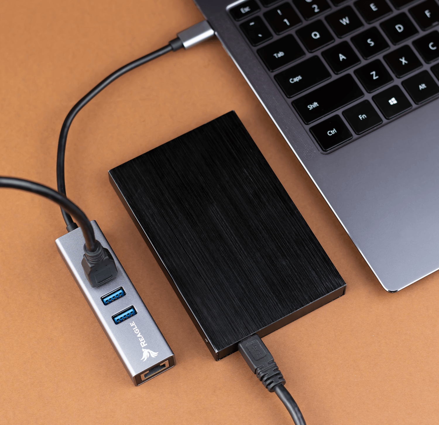 Silberner USB-Hub und eine schwarze externe Festplatte, die mit einem Laptop auf brauner Oberfläche verbunden sind.