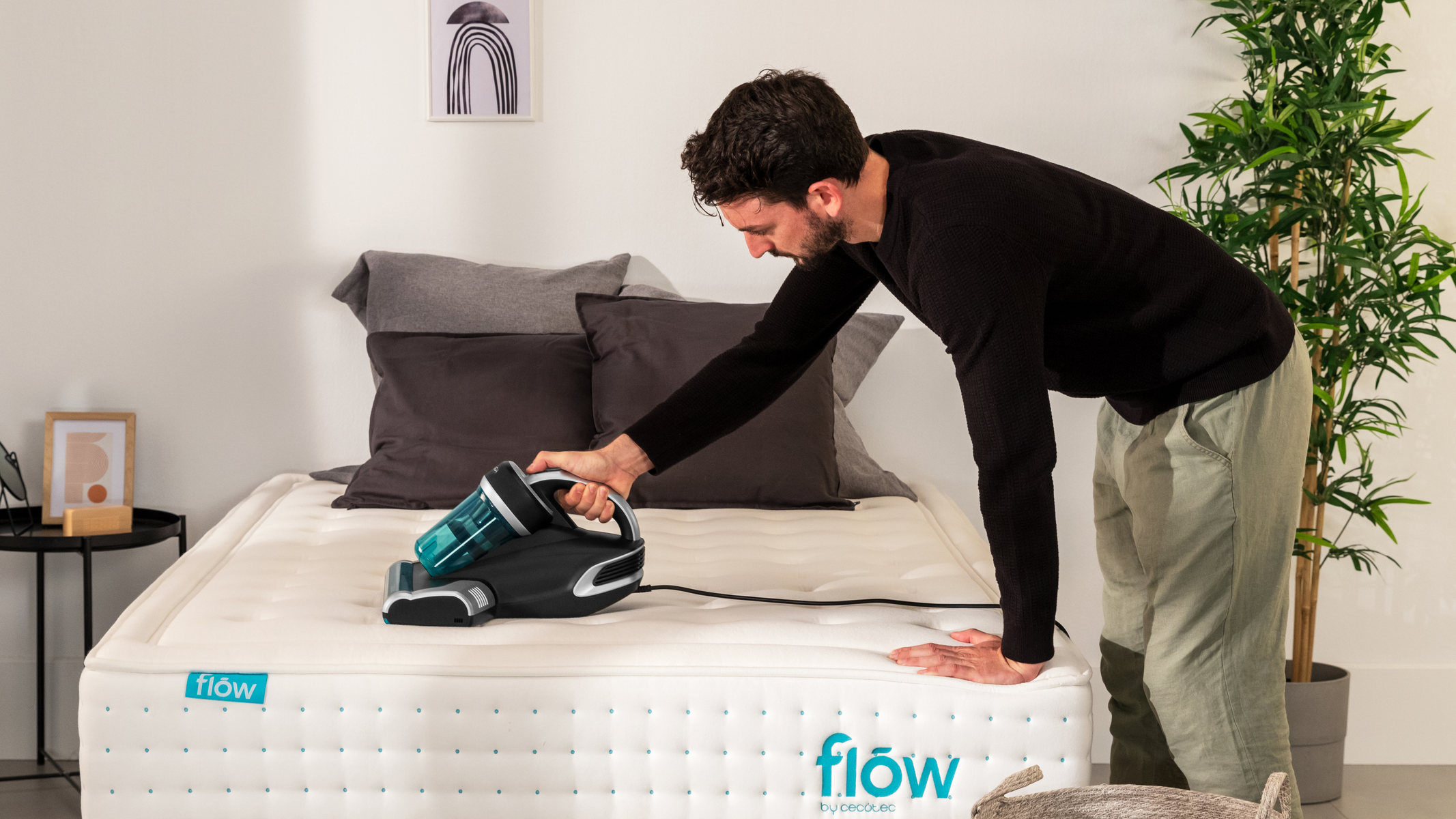 Un uomo aspira un materasso bianco con un aspirapolvere portatile. Finestra e piante sullo sfondo.