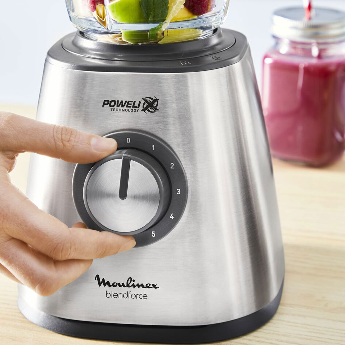 Eine Hand dreht einen Mixer-Regler. Der Mixer ist silberfarben und das Glas enthält Früchte.