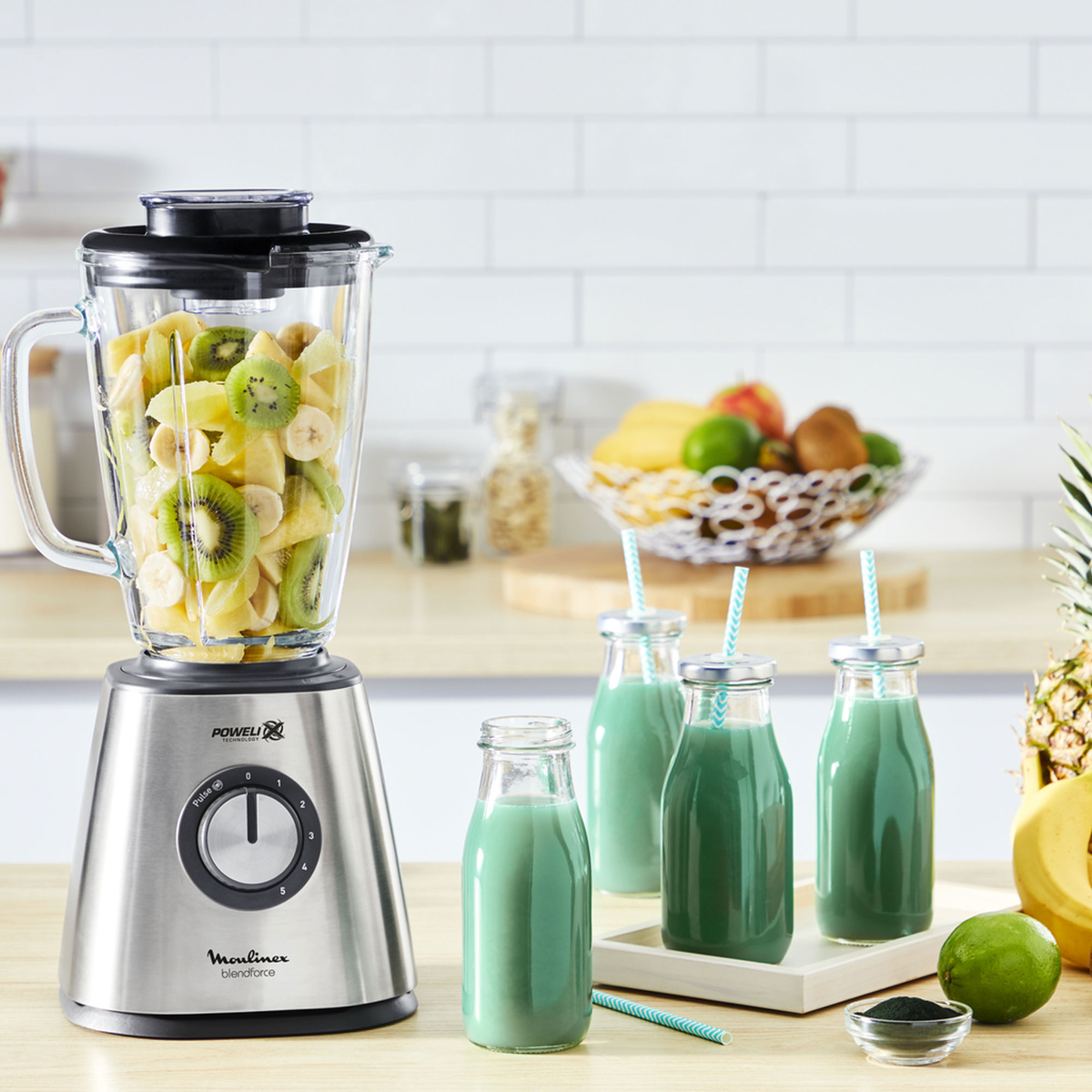 Ein Mixer mit Früchten und Flaschen grünen Smoothies. Küchenszene, gut beleuchtet.