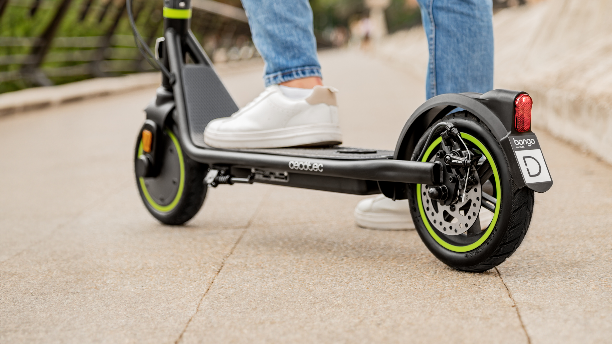 Osoba jedzie czarno-zieloną hulajnogą elektryczną po utwardzonej drodze. Hulajnoga ma logo Bongo.