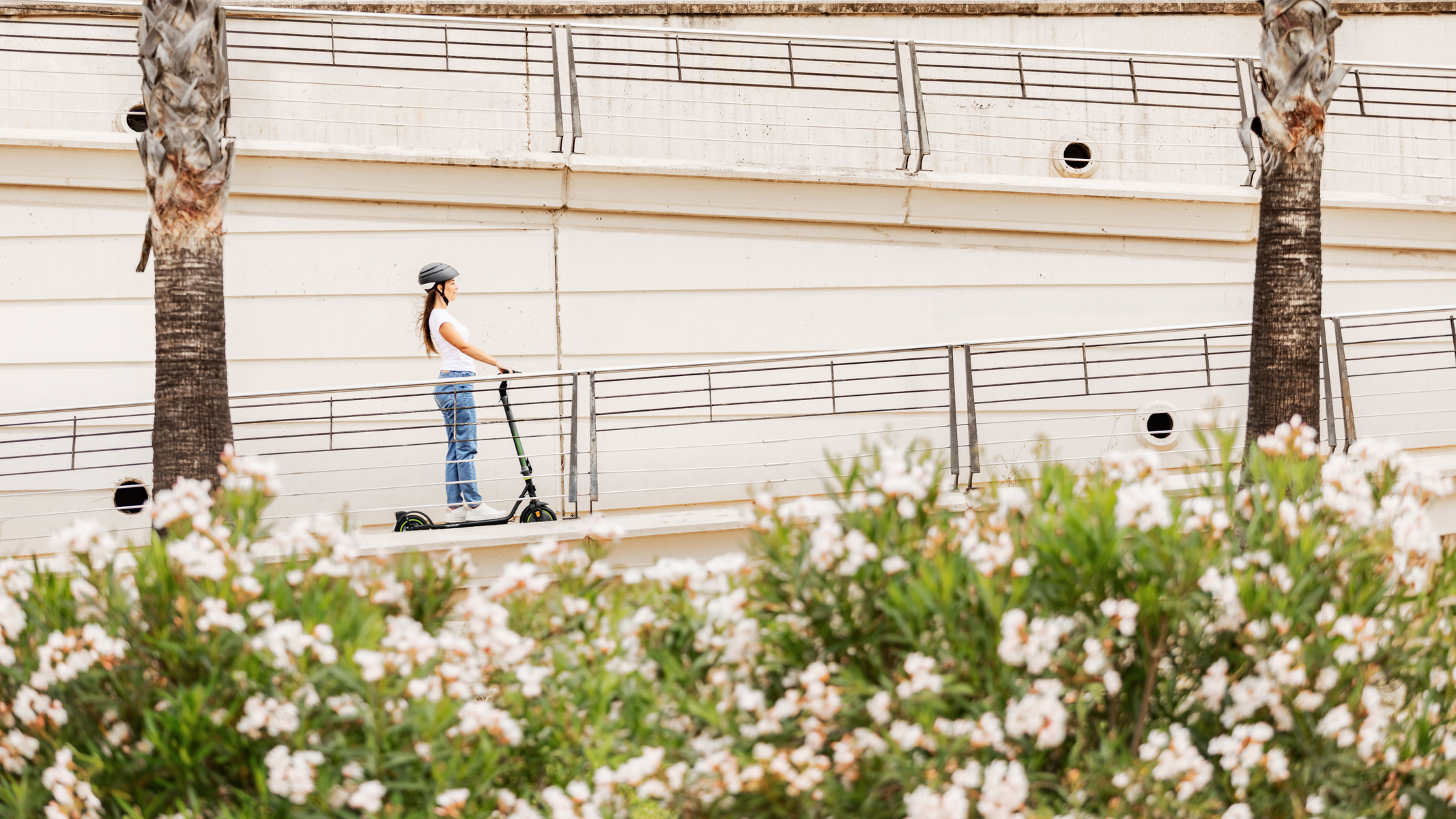Kobieta jadąca zieloną hulajnogą po rampie, z palmami i białymi kwiatami na pierwszym planie.