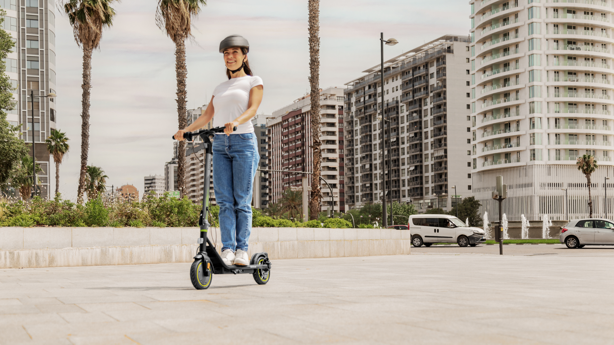 Kobieta jedzie na hulajnodze elektrycznej po utwardzonej ścieżce. W tle budynki i drzewa.