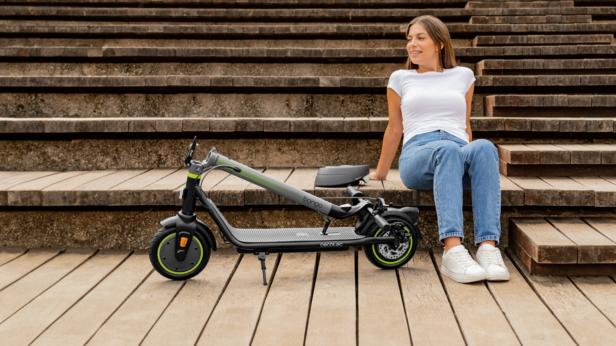 Kobieta siedzi na drewnianych schodach, opierając się o skuter. Uśmiecha się, ma na sobie dżinsy i biały top.
