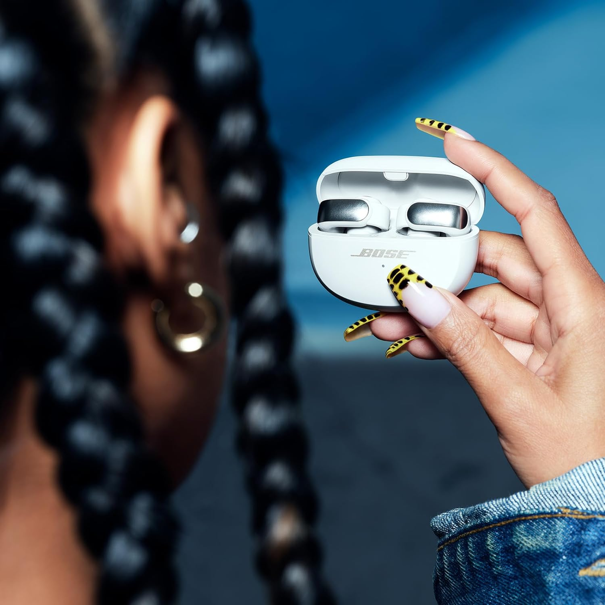 Una persona sosteniendo un estuche blanco de auriculares Bose. El fondo muestra las trenzas de una persona y un degradado azul.