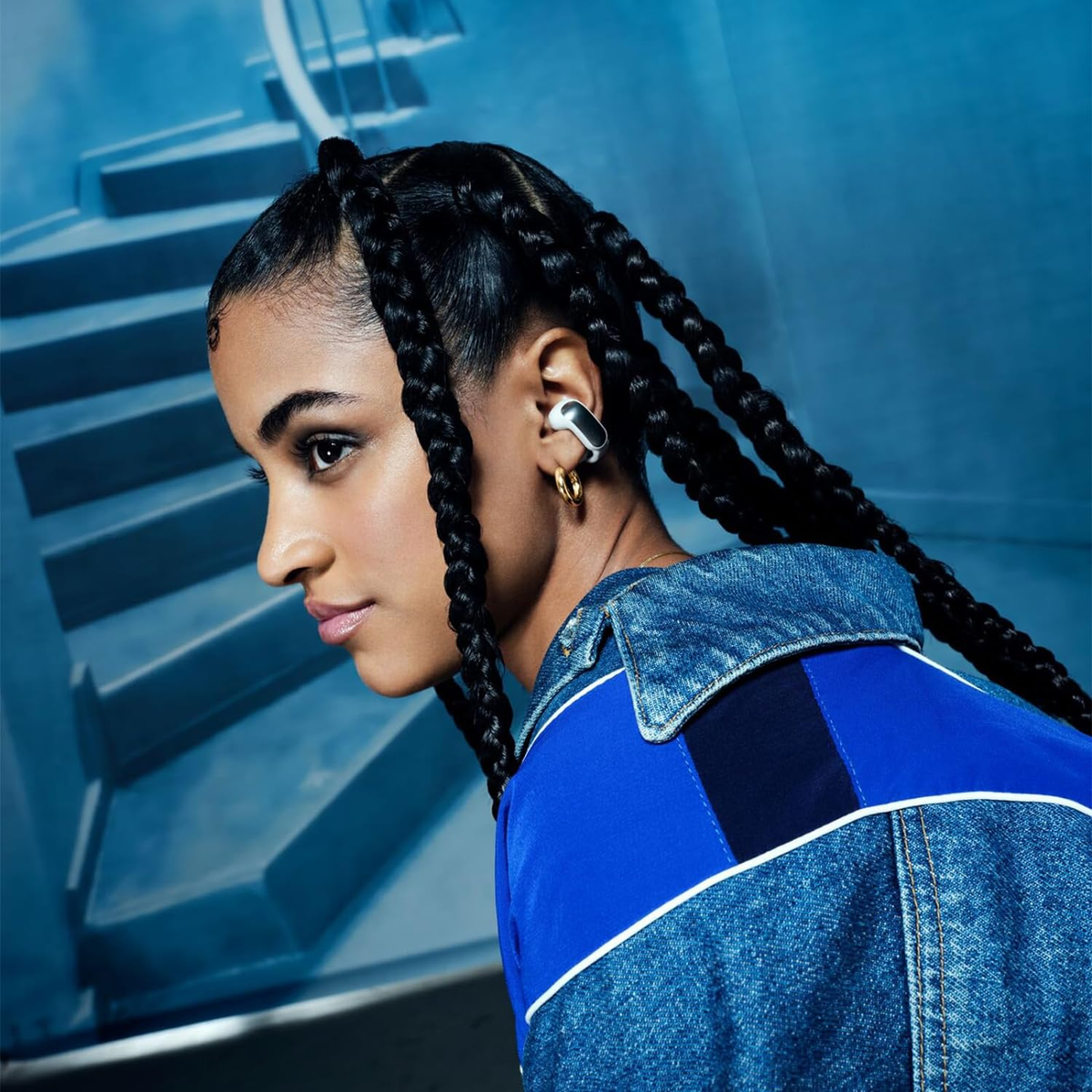 Mujer con trenzas, aretes y una chaqueta vaquera azul. Fondo azul.