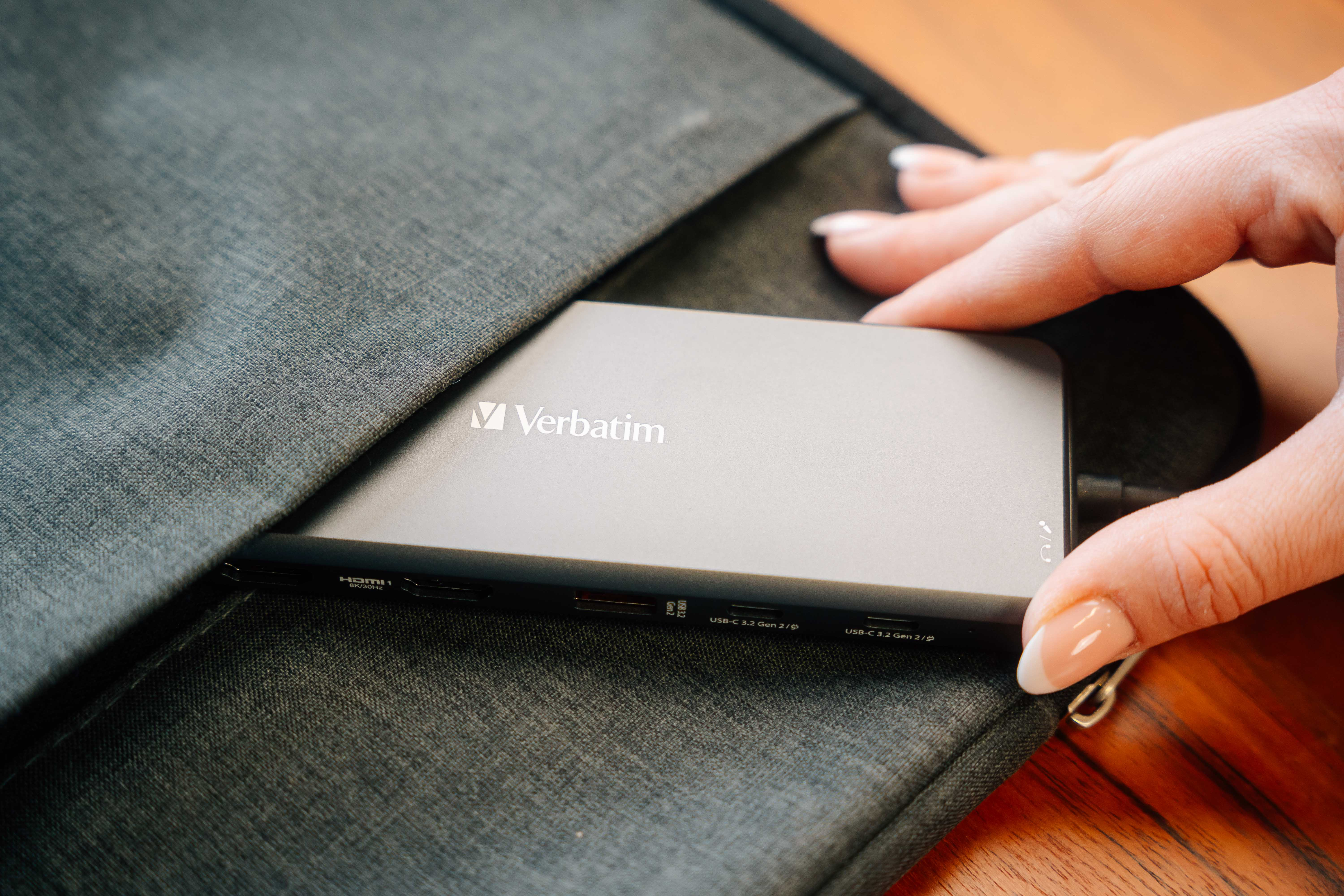 Hand hält eine externe Verbatim-Festplatte aus einer grauen Laptoptasche. Die Festplatte ist silbern.