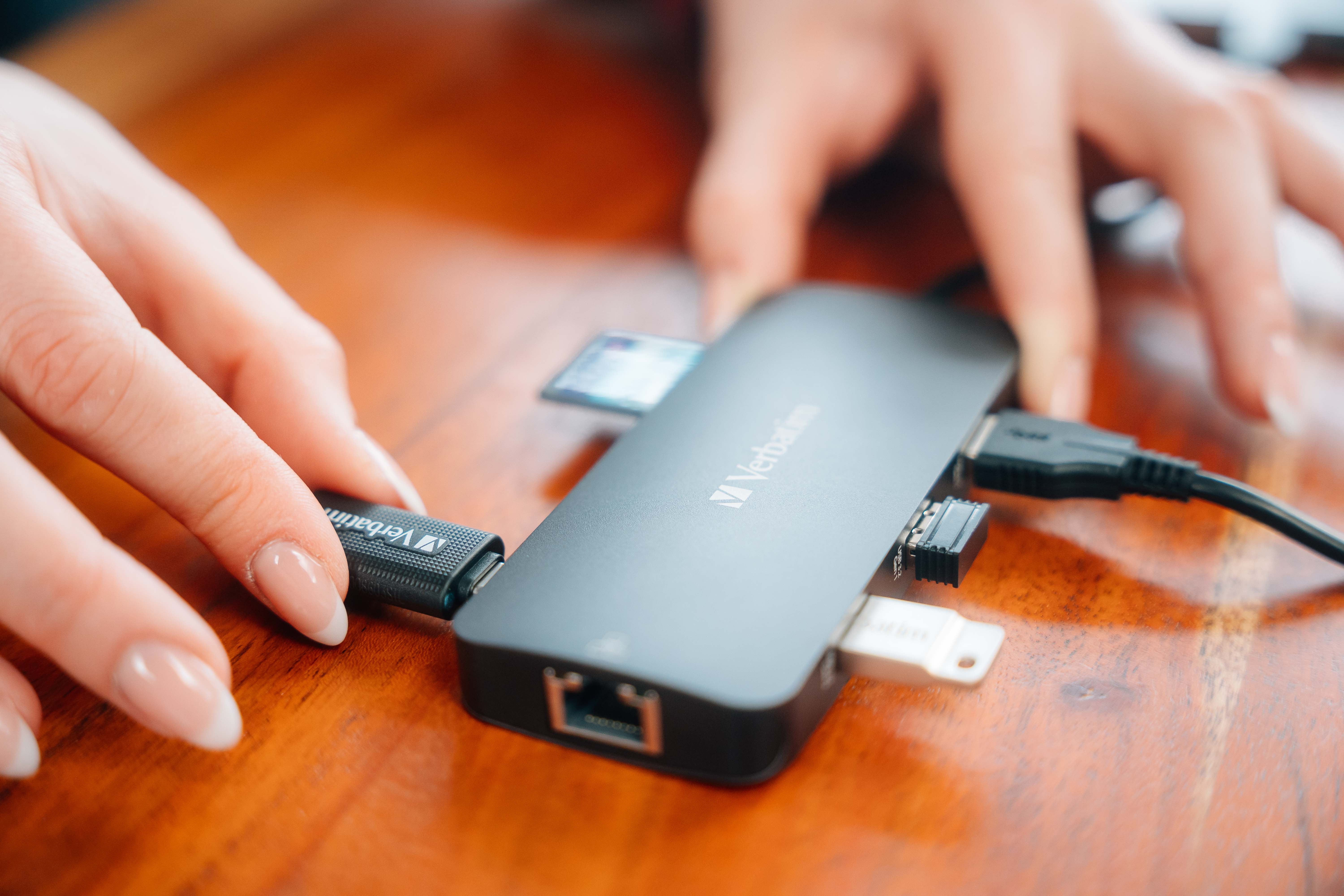 Mani che collegano un cavo USB-C a un hub nero con slot USB e schede. Tavolo di legno.