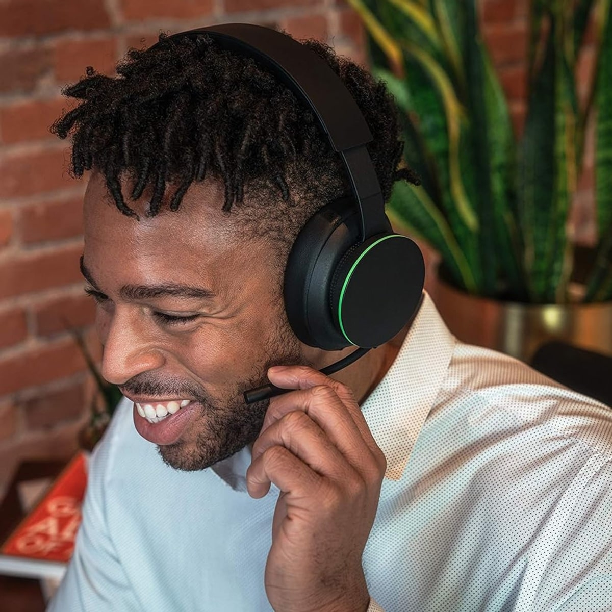 Uśmiechnięty czarny mężczyzna nosi czarny zestaw słuchawkowy Xbox z mikrofonem. Jest w pokoju z rośliną.