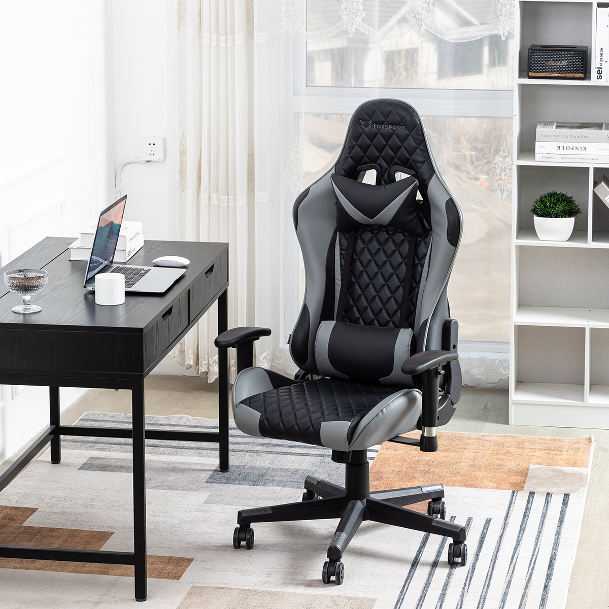 Una silla gaming negra y gris en una habitación. Hay un escritorio con un portátil y una ventana al fondo.