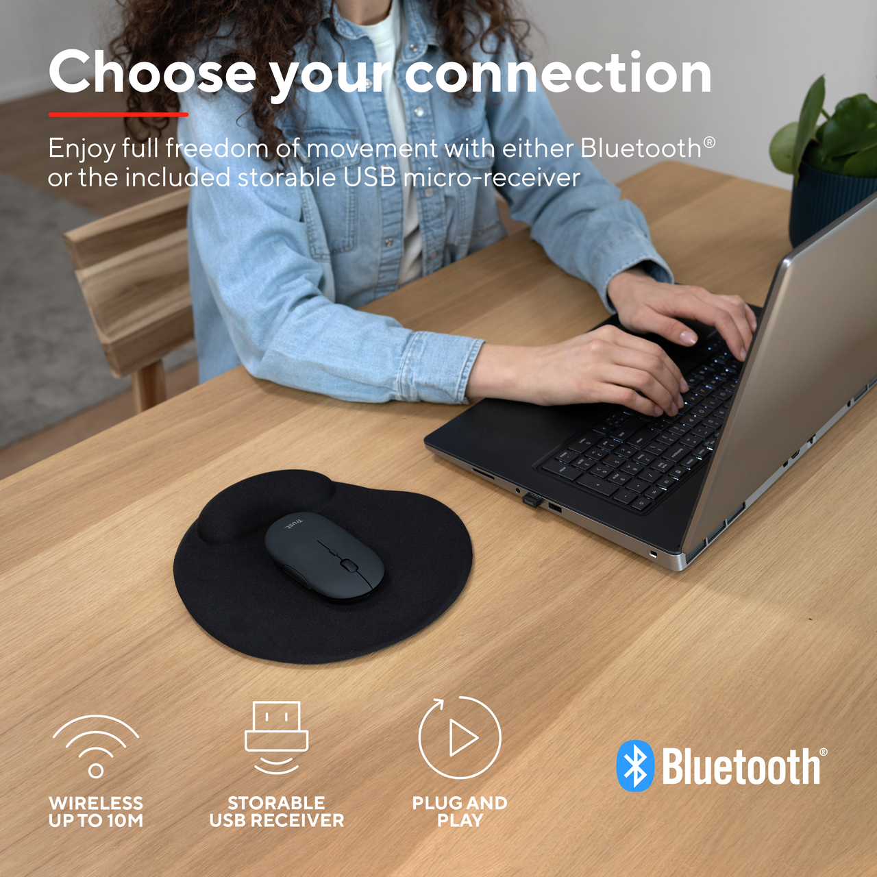 Osoba korzysta z laptopa z myszą i podkładką na drewnianym stole, z ikonami Bluetooth i USB.