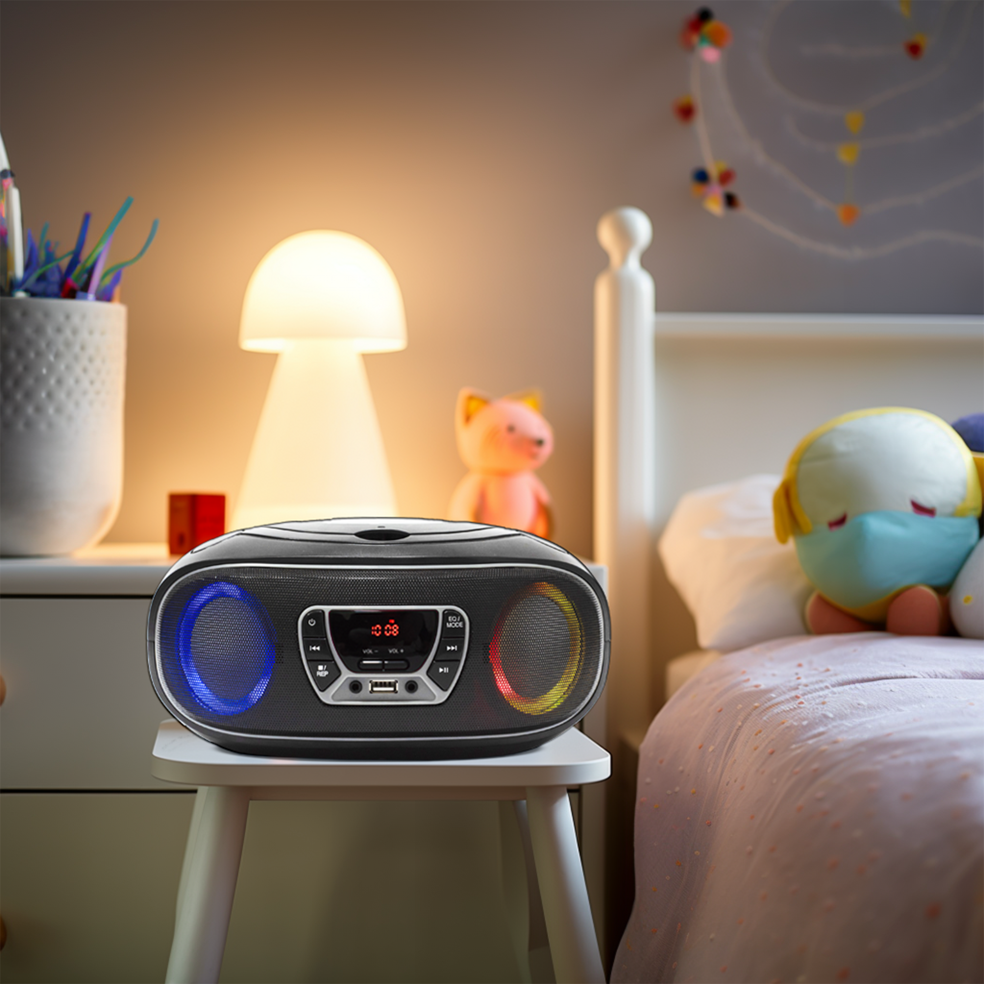 Czarny boombox na białym stołku. Ma kolorowe światła. W tle łóżko i szafka nocna.