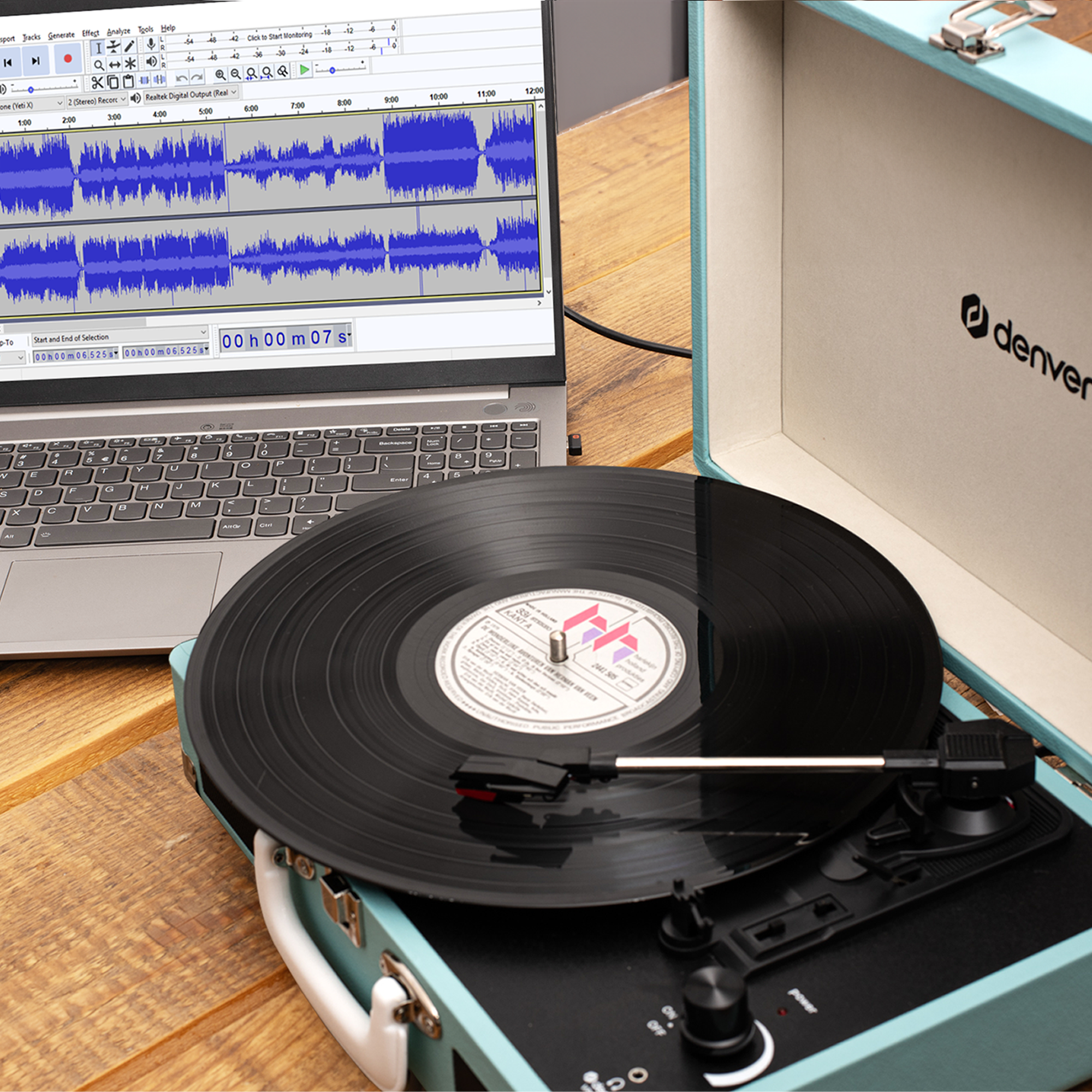 Tocadiscos reproduciendo un disco, laptop mostrando onda de audio, en estuche azul claro, sobre superficie de madera.