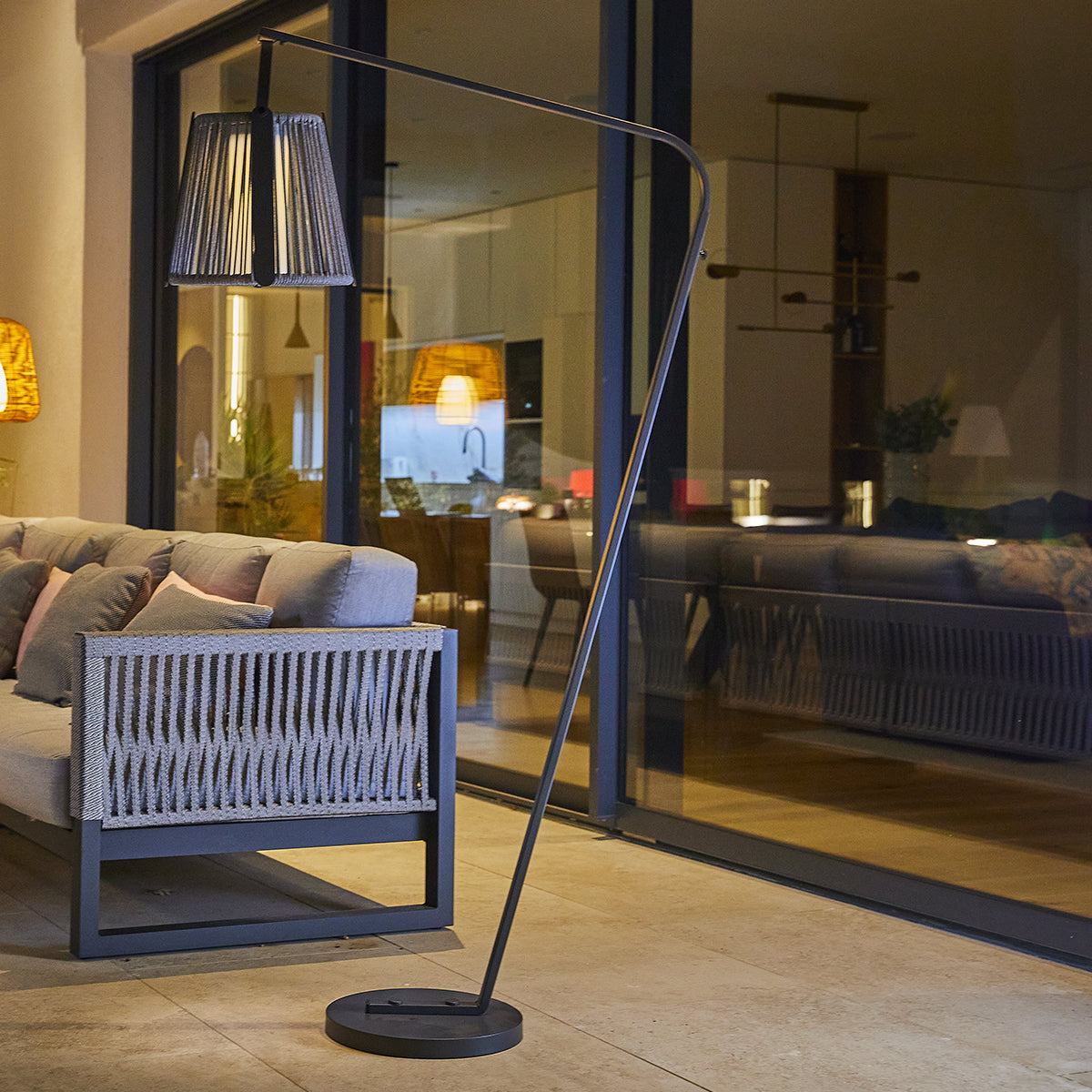 Eine Stehlampe mit grauem Schirm steht neben einem grauen Sofa auf einer Terrasse mit Blick auf ein Haus.
