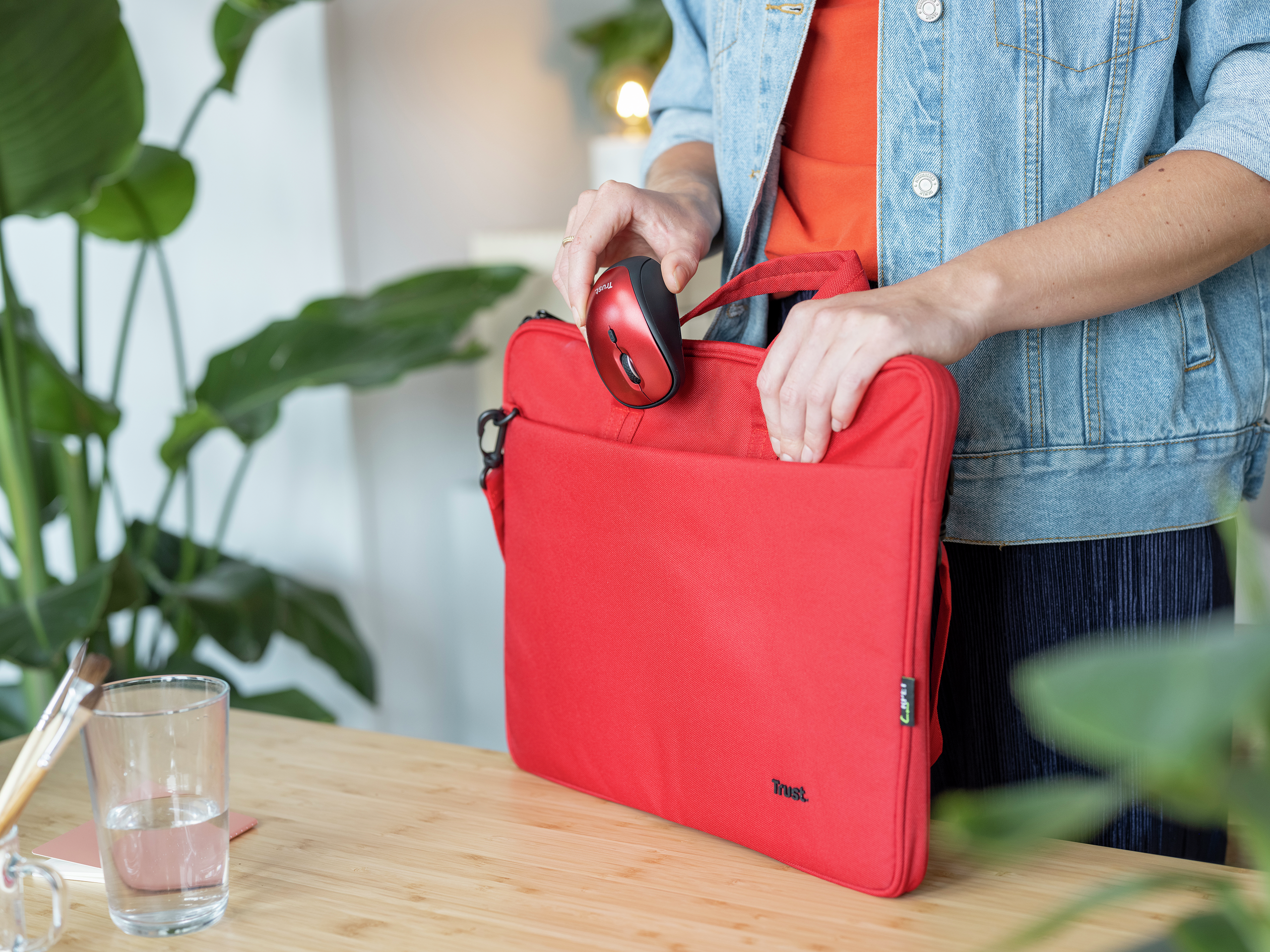 Kobieta wkłada czerwoną mysz do czerwonej torby na laptopa, obok szklanki wody.
