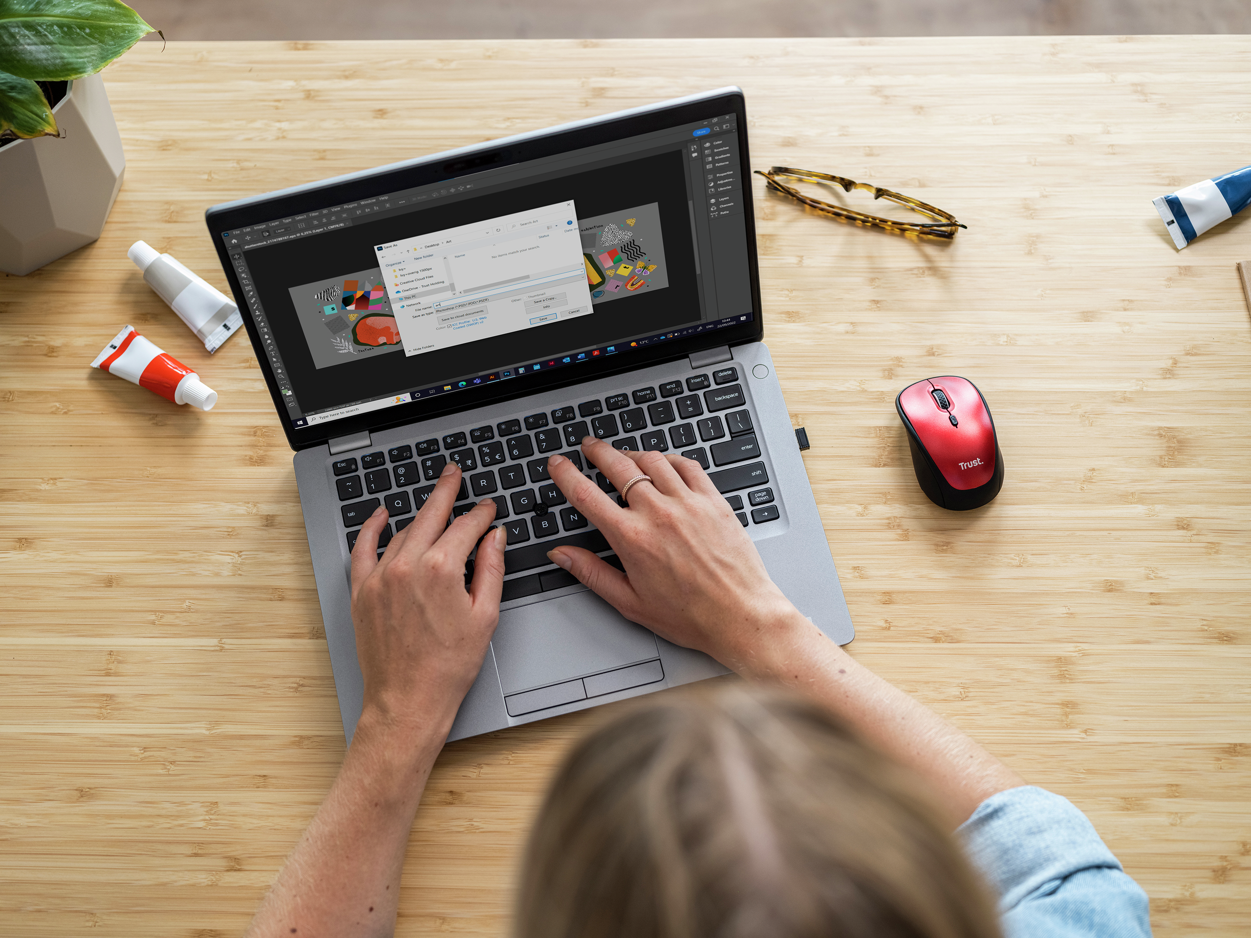 Osoba pisze na laptopie. Akcesoria obejmują czerwoną mysz, okulary i tubki farby na drewnianym biurku.