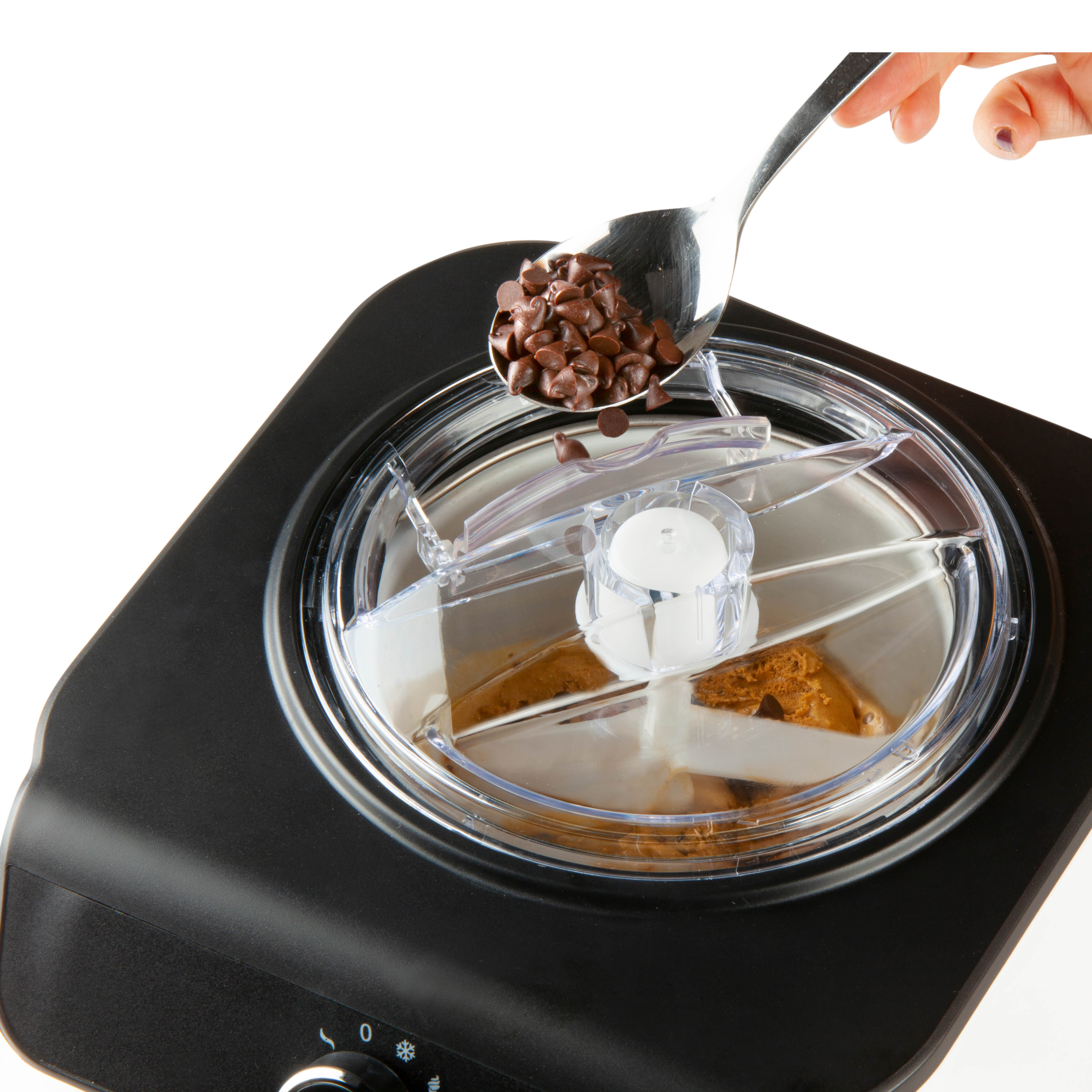 Hand fügt Schokochips in eine Eismaschine. Schwarze Maschine, klarer Deckel, silberner Löffel.